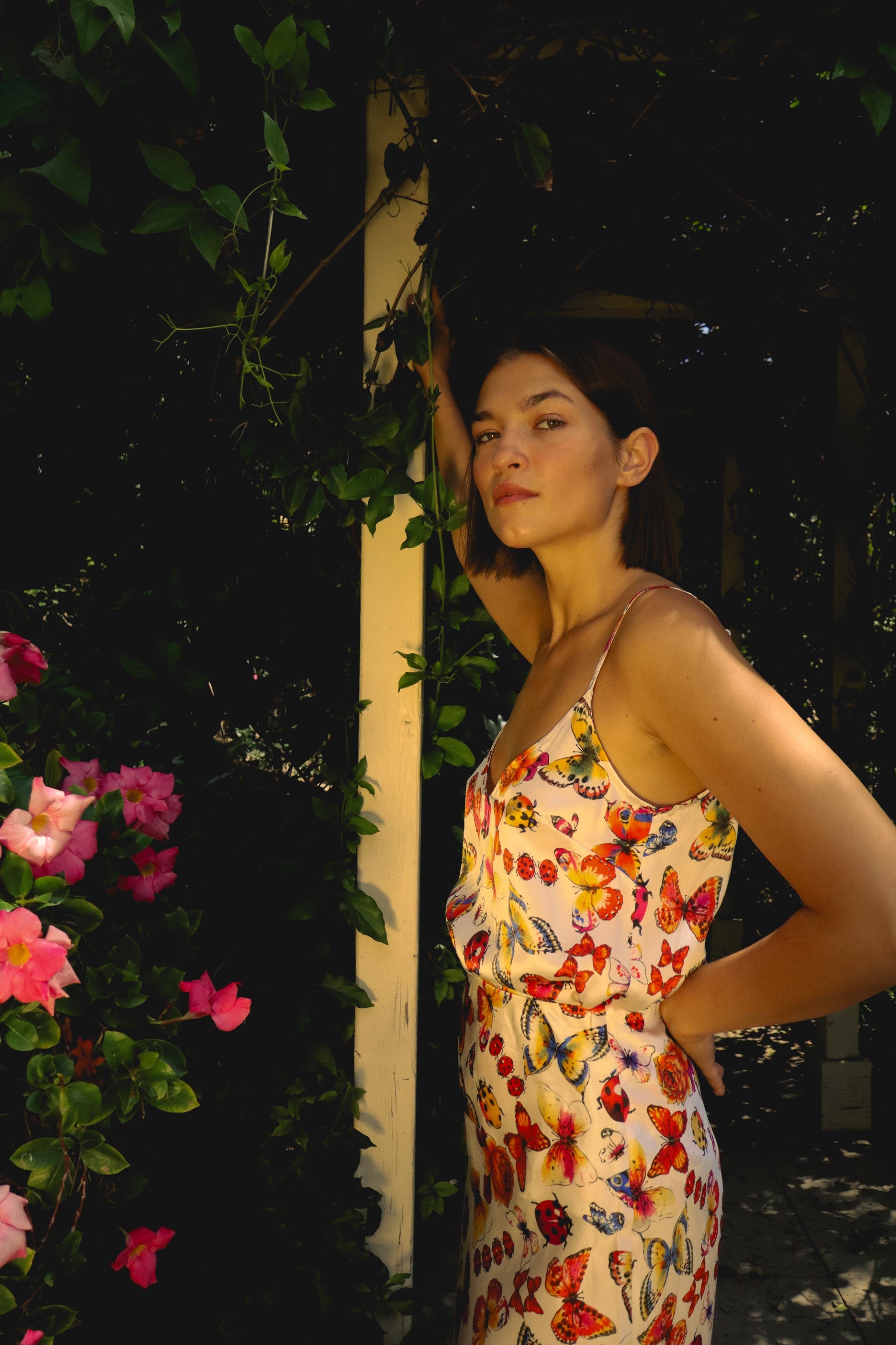 A woman standing outdoors near a trellis adorned with green foliage and pink flowers. She is wearing a Catherine Gee Franka Cami - Ladybug, a sleeveless cami with thin straps featuring a vibrant butterfly print. The colorful floral and butterfly pattern contrasts beautifully with the natural background, making the cami stand out as a stylish and charming piece perfect for a summery, fresh look.