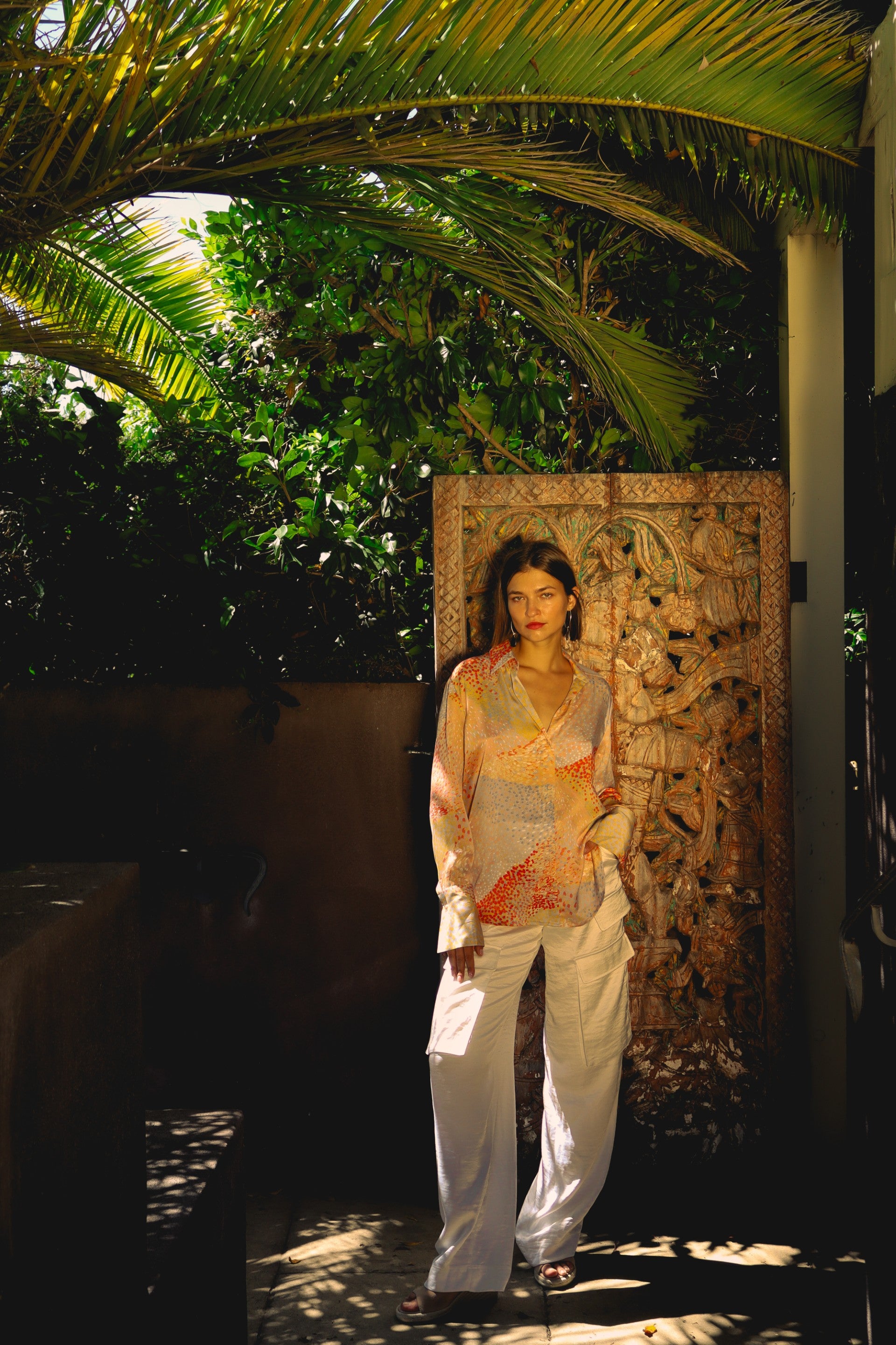 A woman stands in a lush, green environment, with palm fronds overhead. She is wearing the Catherine Gee Daria French Cuff Silk Warm Cosmos Blouse, which features a colorful, abstract pattern in warm tones, paired with white pants. The background includes a decorative carved panel and natural light filtering through the palm tree leaves, creating a mix of shadows that adds an artistic element and enhances the serene ambiance of the scene.