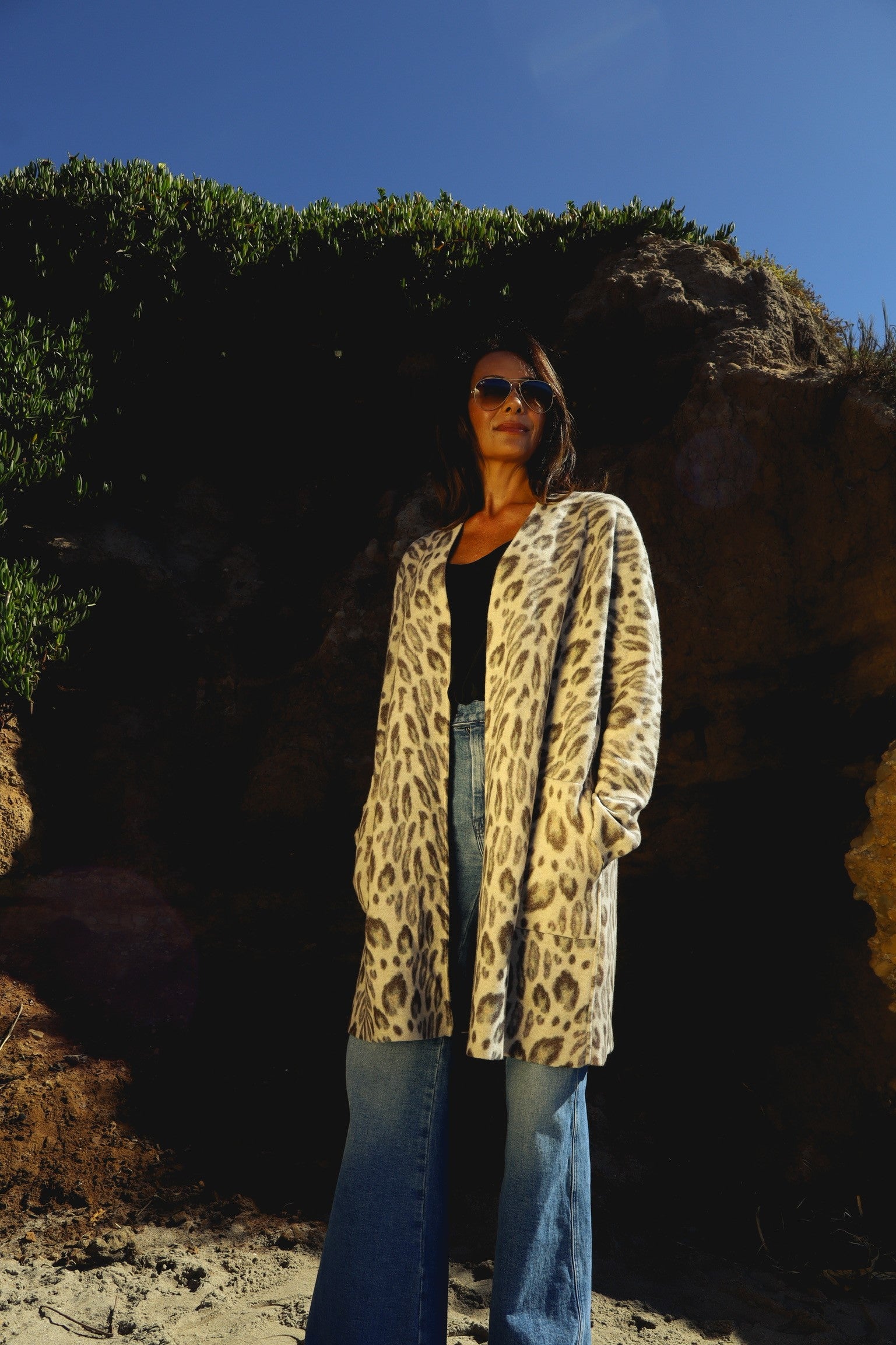 A woman standing outdoors against a backdrop of greenery and rocky terrain, wearing a Catherine Gee Double Face Cashmere Reversible New Leopard coat paired with a black top and blue jeans, with hands in the coat pockets features a sophisticated leopard print design and appears to be made of soft, luxurious cashmere. This stylish and comfortable look is perfect for cooler weather.