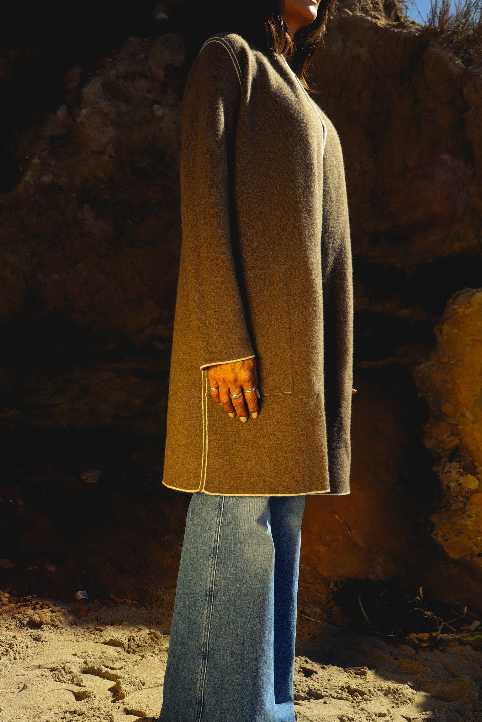 A woman wearing a Catherine Gee Double Face Cashmere Reversible New Leopard Coat. This elegant coat features a neutral color, likely beige or light brown, with a smooth, luxurious texture paired with wide-legged blue jeans, creating a stylish, casual look. The minimalist design with clean lines makes it a versatile and sophisticated outerwear choice. The outdoor setting, with a rocky background and sandy ground, enhances the natural elegance of the coat.