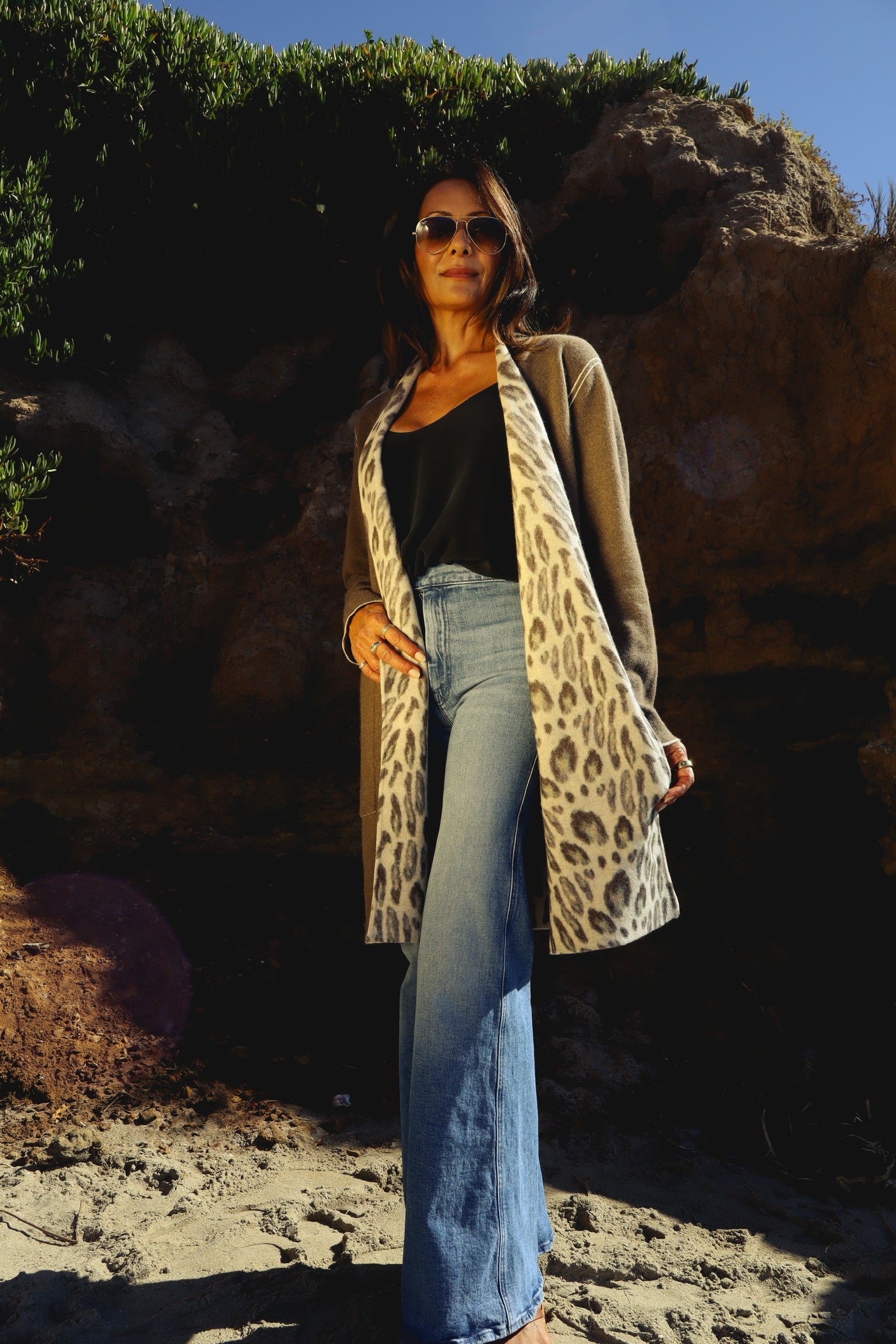 A woman standing outdoors, wearing the Catherine Gee Double Face Cashmere Reversible New Leopard Coat. This luxurious coat features a leopard print on one side and a solid color on the other, allowing for versatile styling paired with a black top and blue jeans, with a natural, sandy, and rocky background highlighting the elegance of the coat. This versatile piece is perfect for adding a touch of sophistication to any outfit.