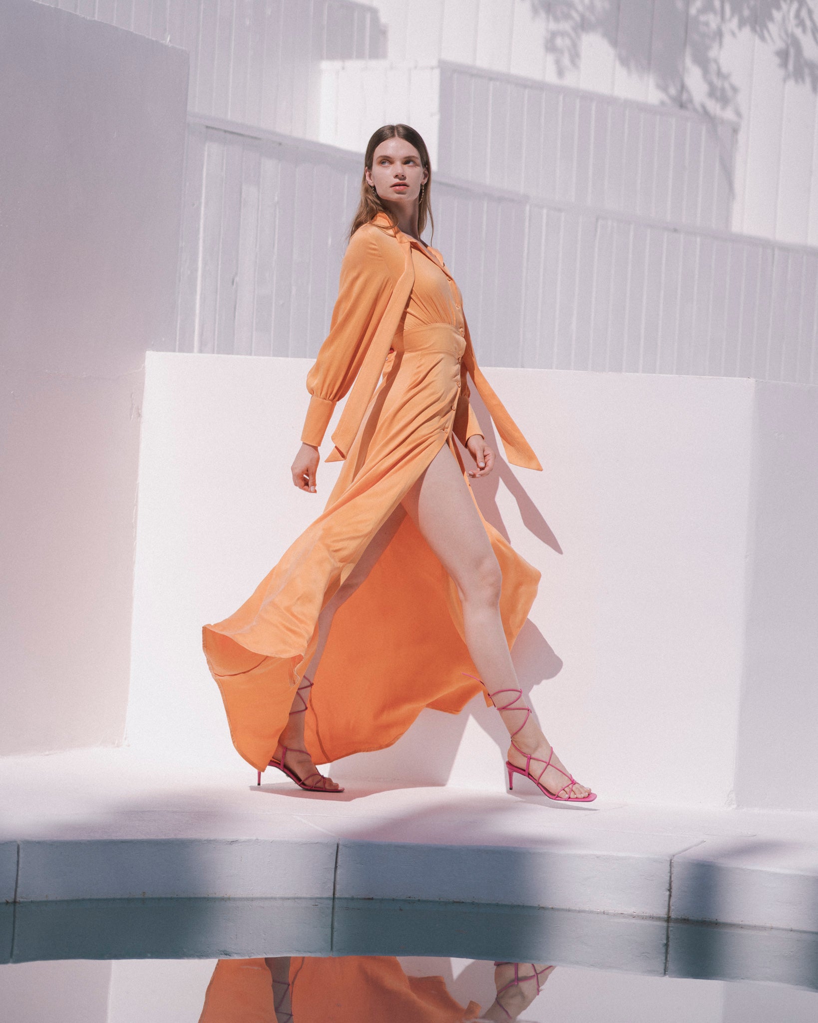 A woman walking confidently in a Catherine Gee Electric Flou High-Slit Long dress is a vibrant Tangerine color with a high slit that reveals one leg paired with strappy high-heeled sandals. The background features white walls and a pool, which reflects the person’s image, adding an artistic touch to the photograph. The flowing fabric and the dynamic pose highlight the elegance and movement of the dress.
