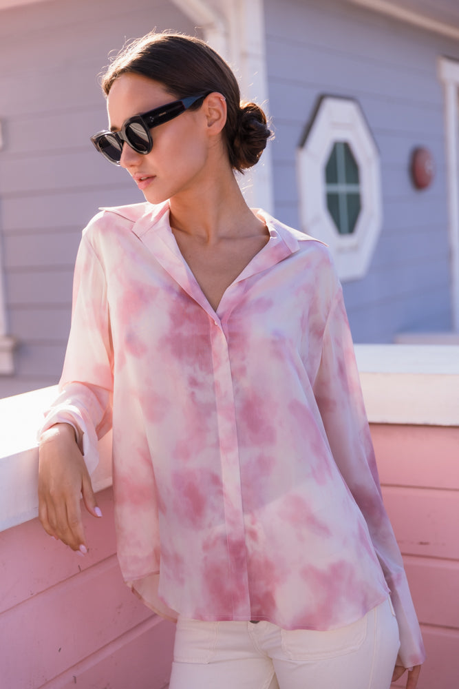 A woman wearing a Catherine Gee Daria French Cuff Silk Pink Dye blouse features a soft, flowing silk fabric with a pink and white tie-dye pattern. It has a relaxed fit with a V-neckline and a classic collar, long sleeves end in elegant French cuffs. She is leaning against a pink railing, with a light blue building in the background that has a white-framed octagonal window. The serene outdoor setting enhances the blouse's stylish and sophisticated appearance, making it a standout piece for any wardrobe.