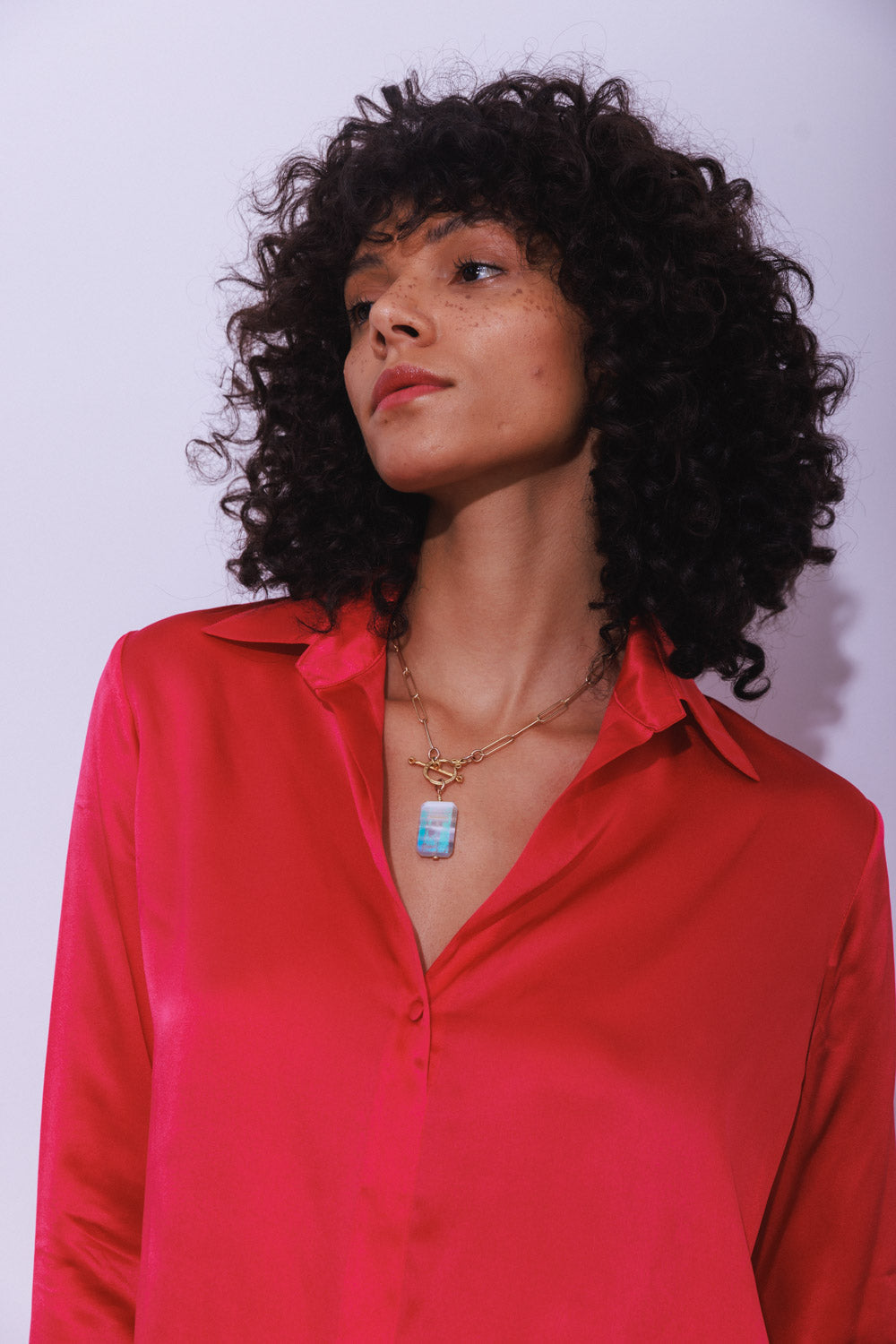 A woman wearing a Catherine Gee Daria French Cuff Silk Blouse - Electric Pink. A bright pink silk blouse with a French cuff design, shiny, smooth texture, and a slightly loose fit. She is also wearing a gold necklace with a rectangular pendant that has a blue and white pattern. The background is plain and light-colored, which makes the vibrant color of the blouse stand out.