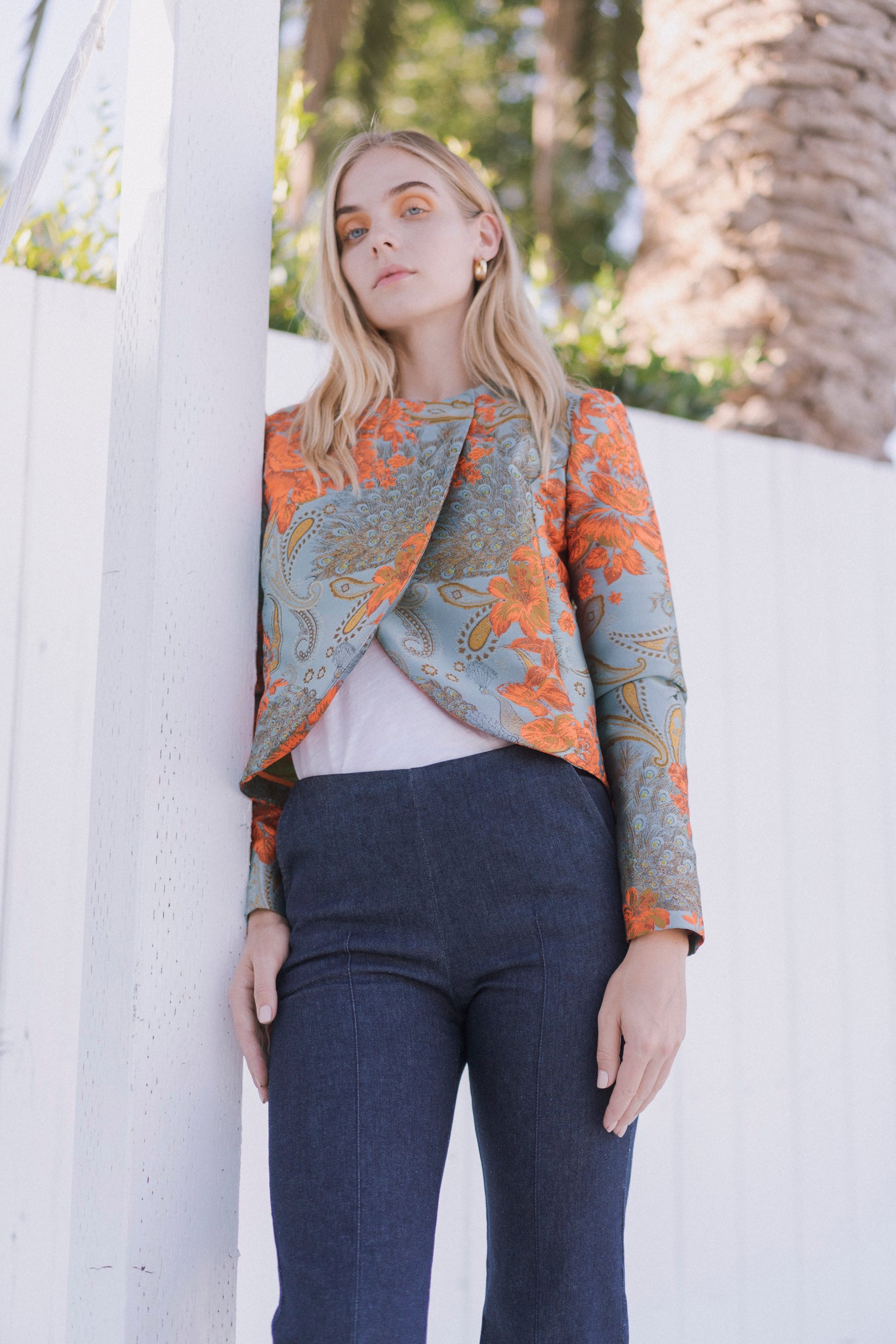 A woman wearing Catherine Gee Emma Tailored Indigo Denim Trousers. These high-waisted, fitted trousers are made from indigo denim fabric, creating a sleek and sophisticated look paired with a colorful, patterned jacket in orange and gray hues over a white top. The outdoor setting includes a white fence and a tree, highlighting the stylish and tailored fit of the trousers.
