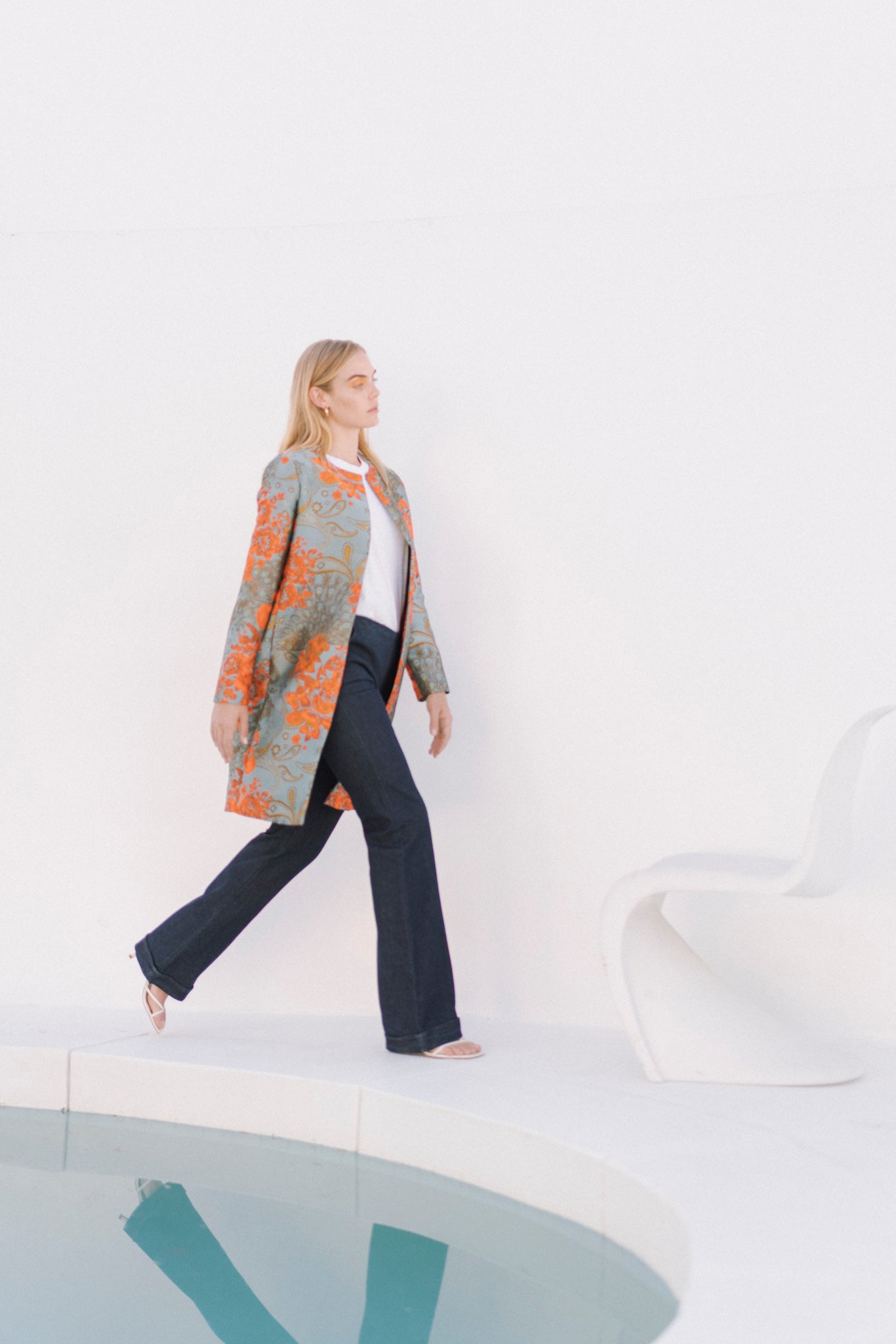 A woman walking beside a pool, wearing the Catherine Gee Emma Tailored Indigo Denim Trouser. These dark blue trousers have a sleek, tailored fit that exudes sophistication paired with a white top and a long, patterned coat featuring orange and grey designs. The minimalist setting, with a white background and a modern white chair, adds to the stylish and contemporary aesthetic of the ensemble.