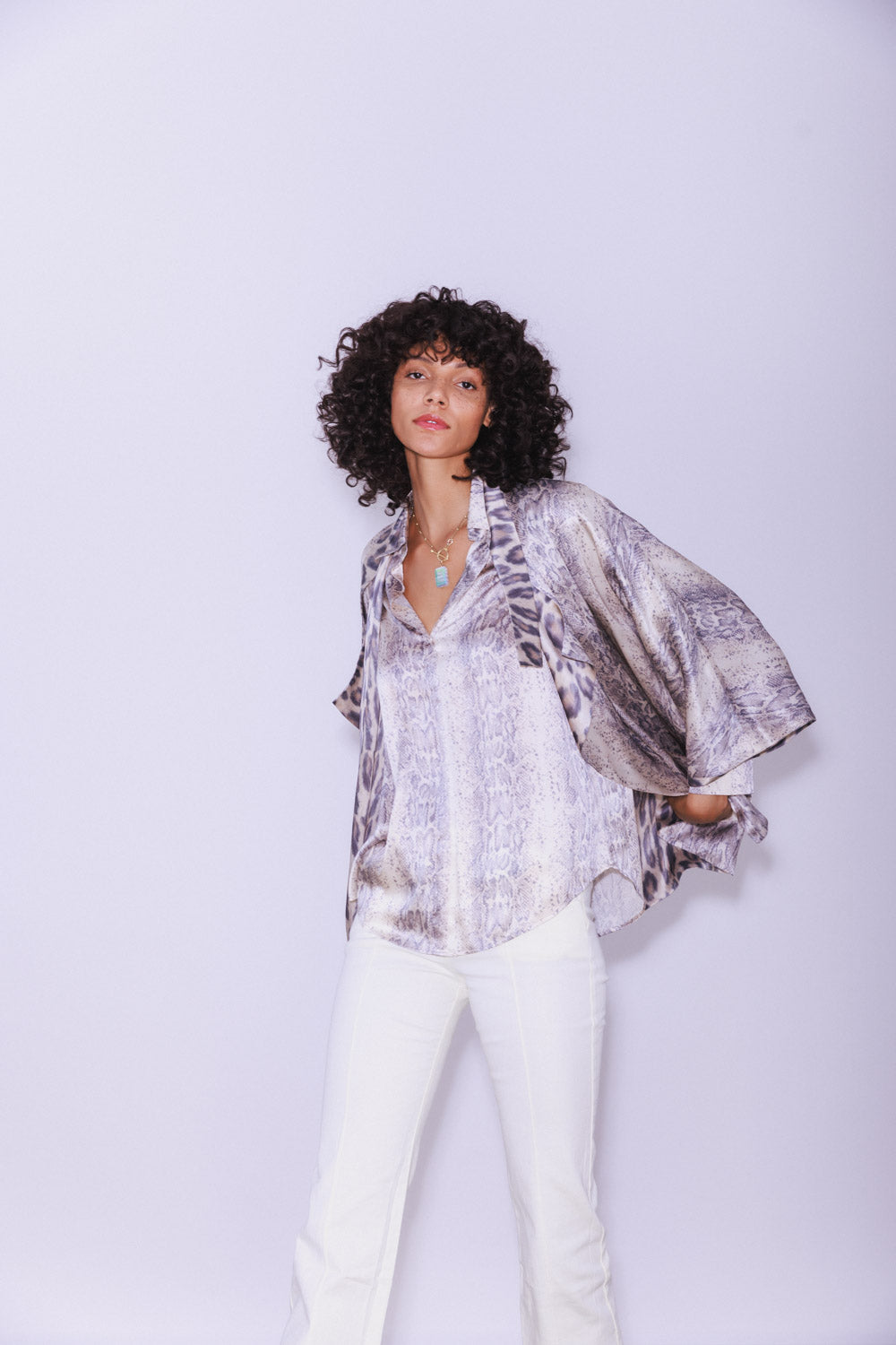A woman wearing a Catherine Gee Daria French Cuff Silk Sedona Snake blouse features a snake print pattern with a loose, flowing fit and wide sleeves that end in French cuffs paired with white pants, on a plain light color background, which highlights the outfit. The blouse's luxurious silk material and unique design make it a standout piece, perfect for a stylish and sophisticated look.
