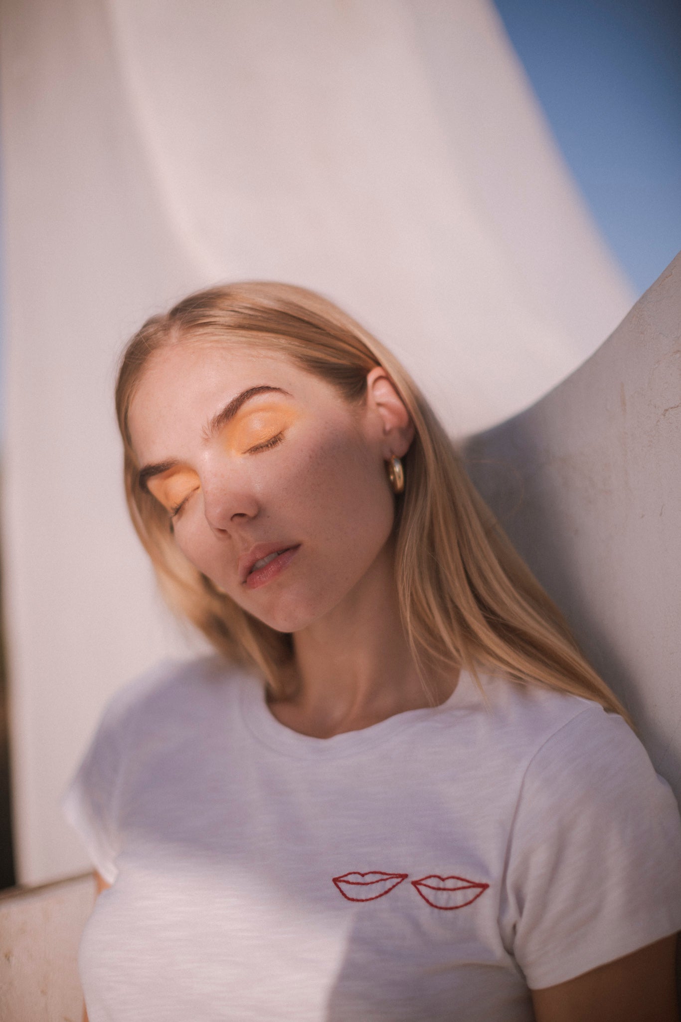 A woman with long blonde hair wears gold hoop earrings wearing a Catherine Gee Embroidered Cotton Lips white t-shirt that features a minimalist design with two red embroidered lips on the left side of the chest. The background appears to be an outdoor setting with a white structure and a clear blue sky highlighting the casual yet stylish nature of the t-shirt, making it interesting for fashion enthusiasts.