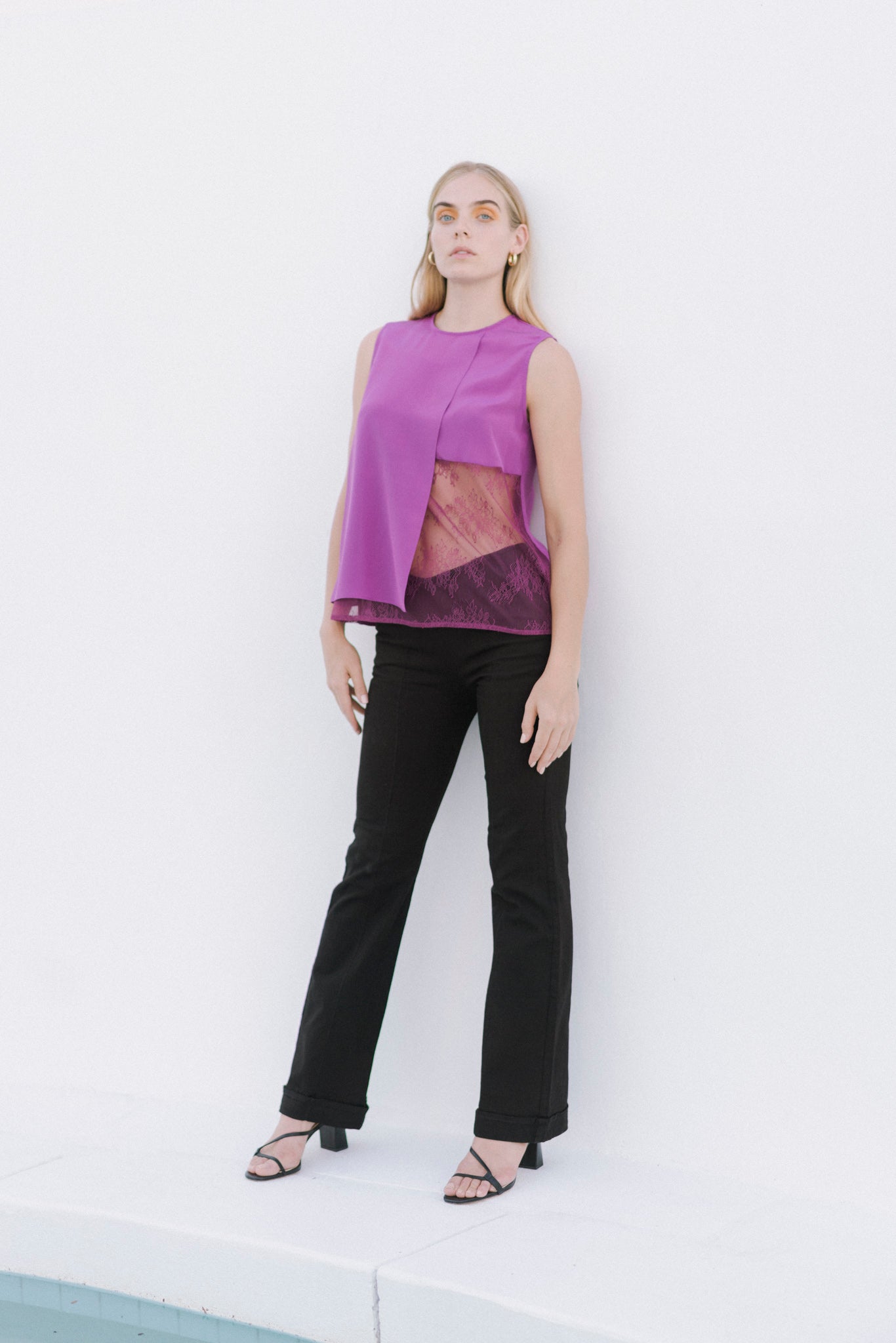 A woman wearing Catherine Gee Emma Tailored Black Twill Denim Trousers. These sleek, high-waisted trousers feature a tailored fit with a slight flare at the bottom paired with a sleeveless purple top that has an asymmetrical design and a sheer section in the middle. The combination creates a stylish and modern look, perfect for both professional and casual settings.