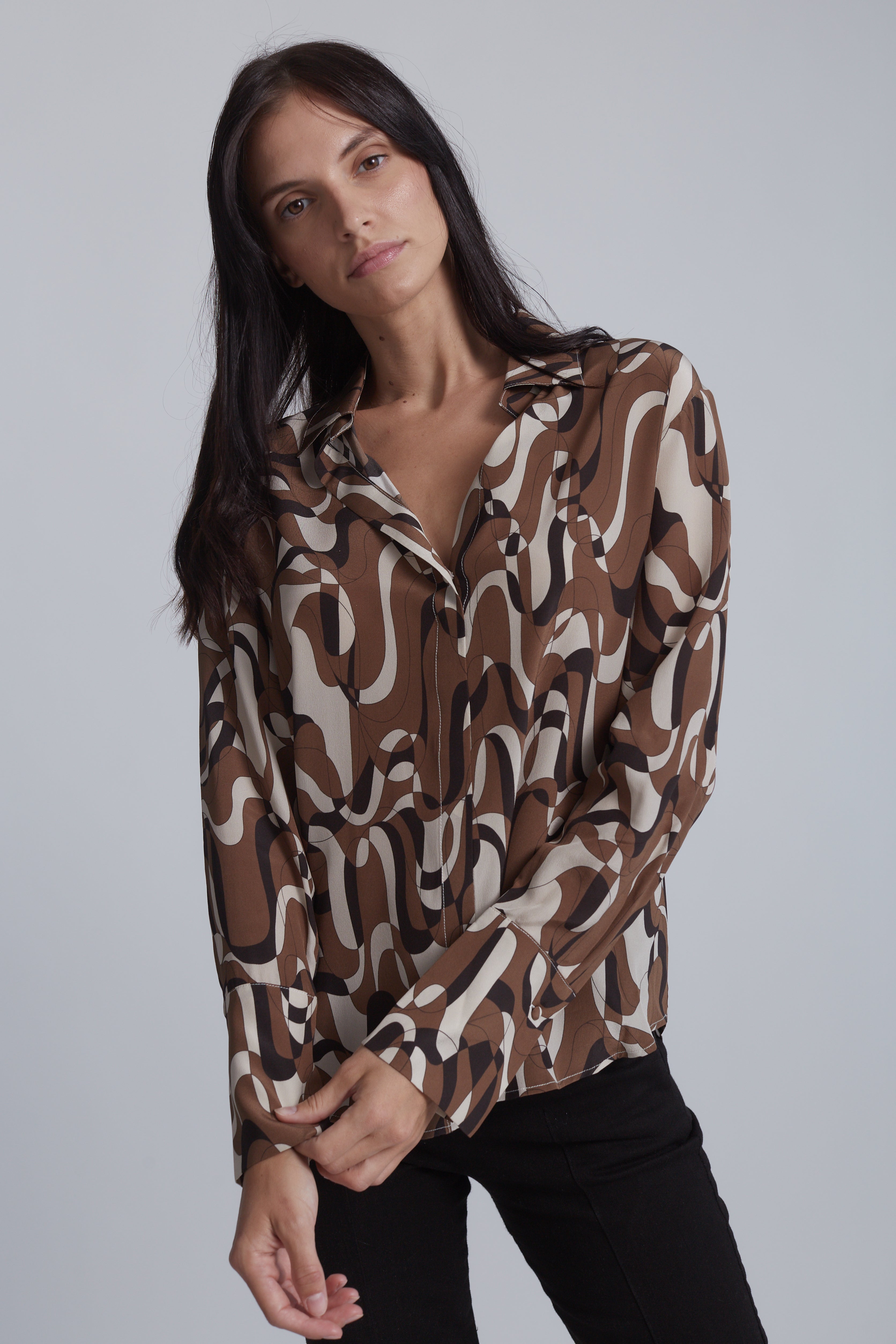A woman wearing a Catherine Gee Daria French Cuff Silk Barcelona blouse features a bold, abstract design with intertwining brown, white, and black shapes. She is standing with her arms slightly crossed and head tilted, showcasing the blouse's relaxed fit and stylish pattern on a plain and neutral background, highlighting the blouse.