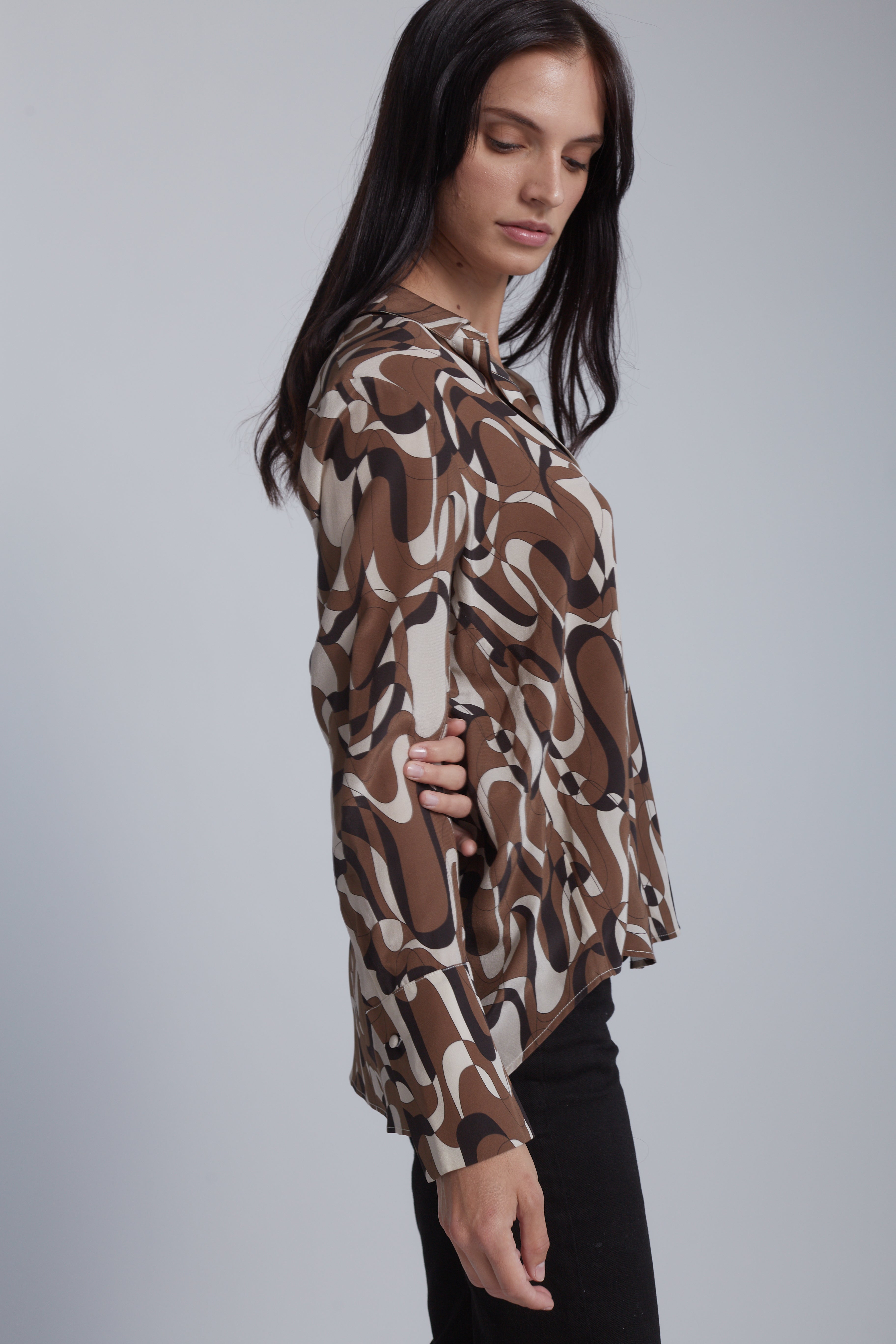 A woman wearing a Catherine Gee Daria French Cuff Silk Barcelona blouse features a bold, abstract design with intertwining brown, white, and black shapes. She is standing sideways with one hand on the hip, showcasing the blouse's fit and design.