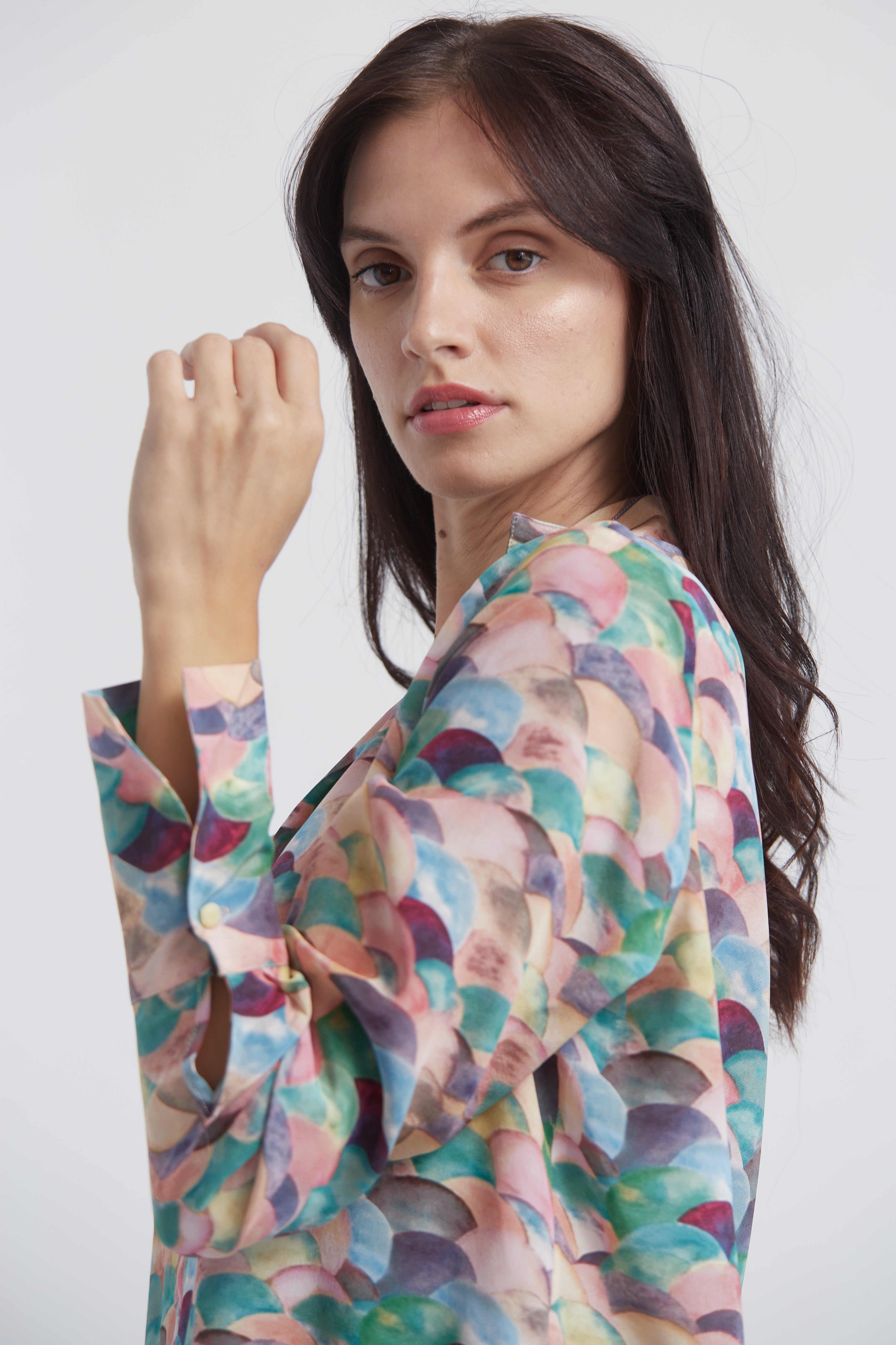 A woman with long, dark hair in side view, wearing a Catherine Gee Daria French Cuff Silk-Lima blouse features a colorful, abstract design with overlapping semi-circles in shades of pink, blue, green, and purple. The blouse has long sleeves with French cuffs, which are fastened with a button. 