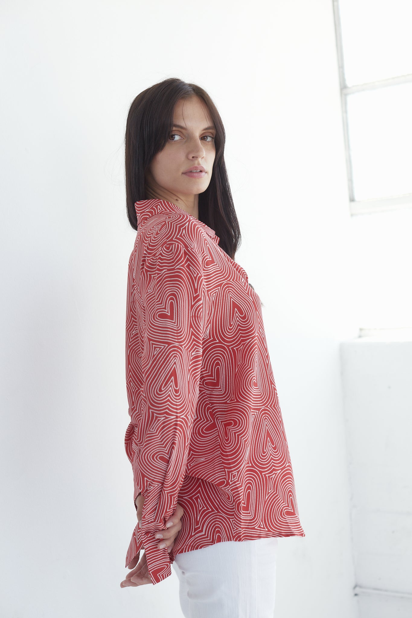Woman wearing a Catherine Gee Daria French Cuff Silk Bowie Heart blouse featuring red with a white heart pattern and has a loose fit. She is standing sideways, with the left arm slightly bent and the right arm hanging down in a minimalistic white room background.