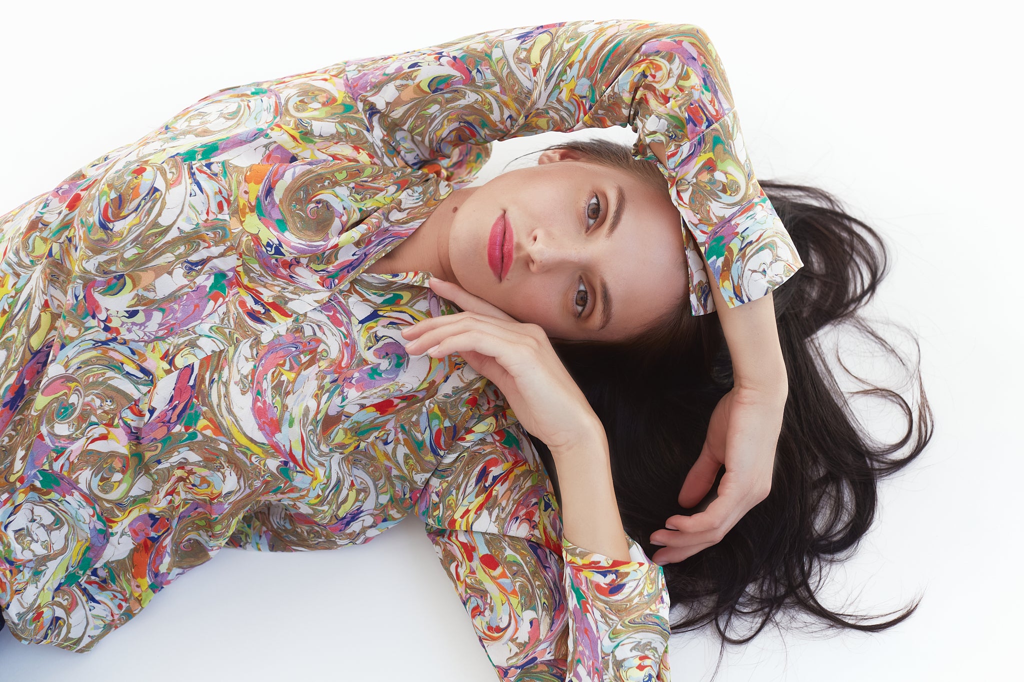 A woman lying down, wearing a Catherine Gee Daria French Cuff Silk Paint Swirl blouse features a vibrant and intricate pattern with swirling designs in various colors, including shades of gold, red, purple, blue, and green. The fabric appears to be smooth and luxurious in texture and has long sleeves with French cuffs, adding a touch of elegance to the overall design, and highlighting the artistic and fashionable aspects of the blouse, making it relevant for those interested in unique and stylish clothing.