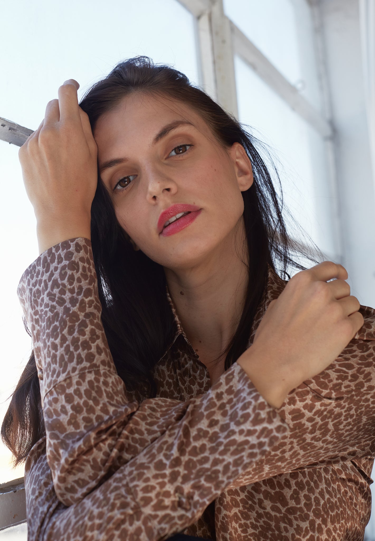 A woman wearing a Catherine Gee Daria French Cuff Silk-Electric Leopard blouse features a leopard print pattern and has French cuffs. She poses with one hand touching her head and the other arm bent on a background showing a window with white frames in natural lighting coming to her back.