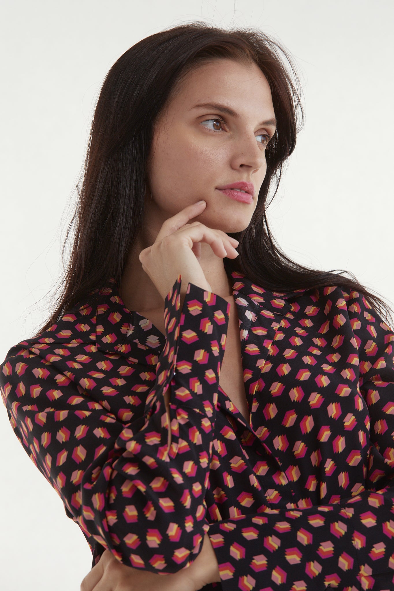 A woman with long, dark hair posing with one hand resting on the chin and the other arm crossed over the body wearing a Catherine Gee Daria French Cuff Silk-Libros Print blouse features a pattern of small, colorful book-like shapes on a dark background.