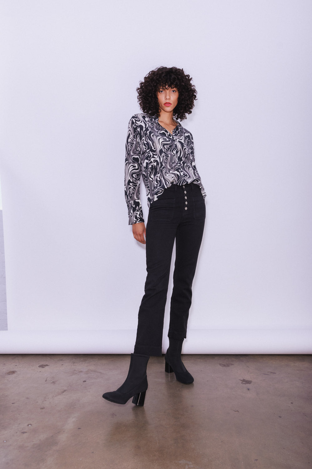 A woman standing against a plain white background, wearing a Catherine Gee Daria French Cuff Silk Blouse Dali features a swirling, abstract black-and-white design paired with high-waisted black pants with a row of buttons on the front and black heeled boots, creating a stylish and modern look.