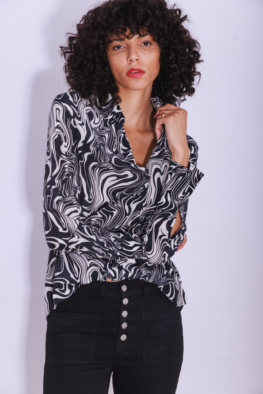 A woman wearing a Catherine Gee Daria French Cuff Silk-Dali blouse features a black and white swirling design and is paired with high-waisted black pants with a row of buttons on a plain white background.
