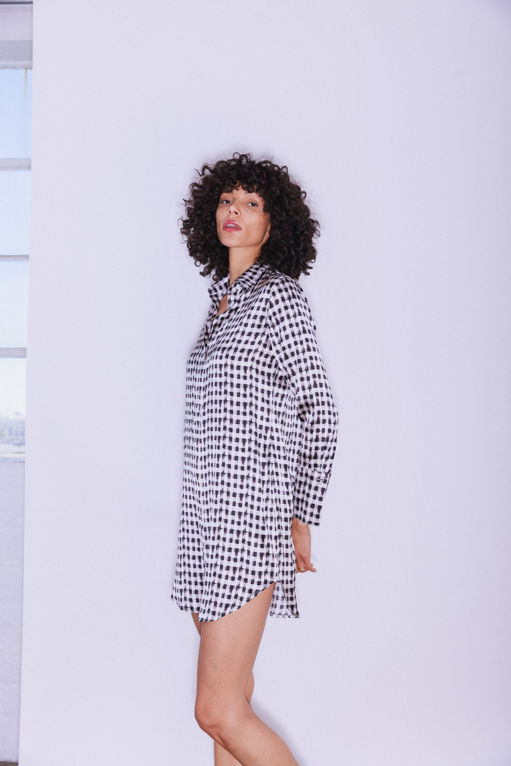 A woman wearing a Catherine Gee Daria Silk Shirt Grosgrain Black and White dress features a striking black and white checkered pattern, long sleeves, and a loose, comfortable fit. The hemline falls above the knees, and the dress includes a button-down front and a classic collar. The plain background emphasizes the bold design of the dress.