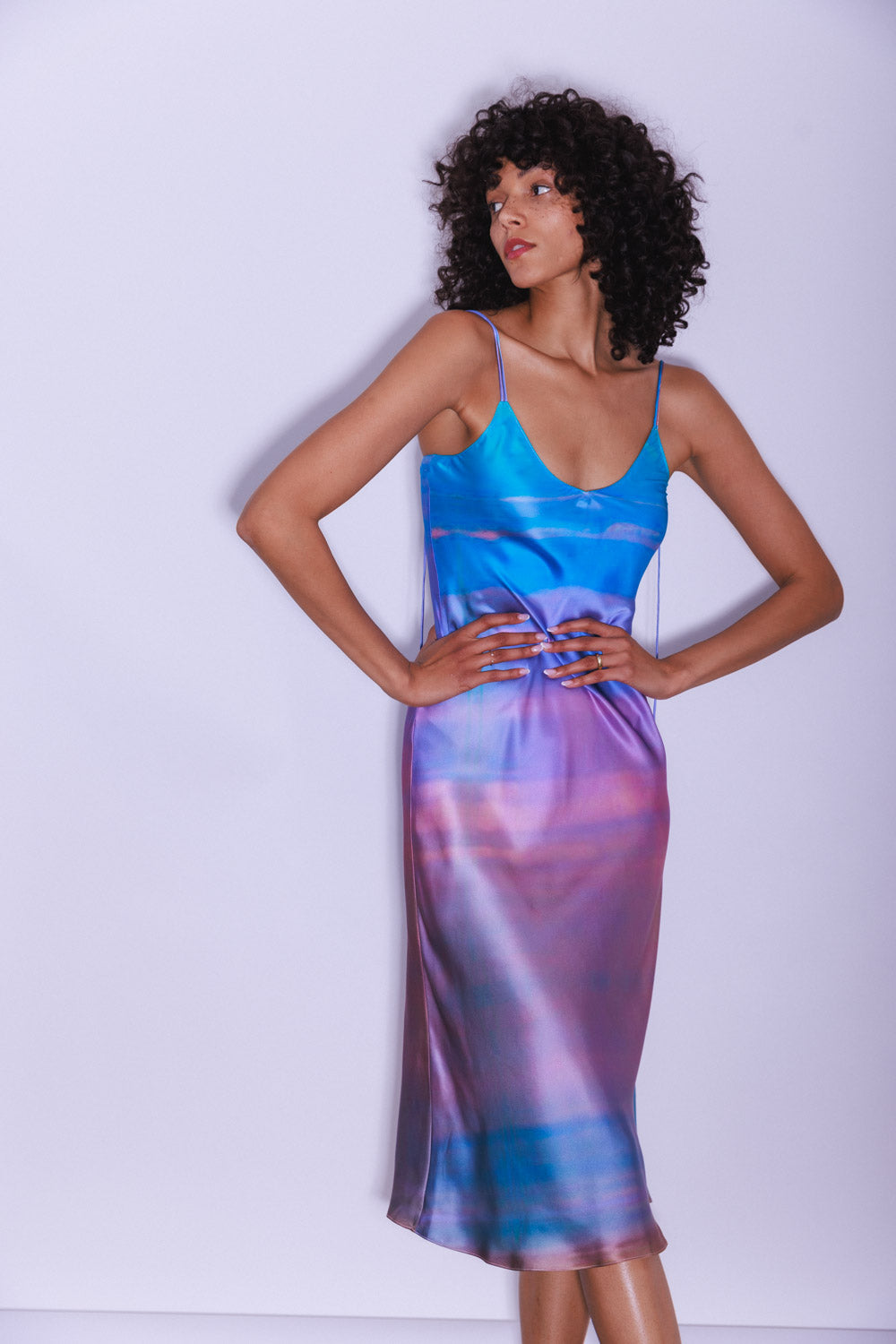A woman standing with hands on hips, facing away from the camera wearing the Catherine Gee Emma Peruvian Quartz Slip Dress. This midi-length dress features a vibrant, multicolored pattern with shades of blue, purple, and pink, has thin straps, and a sleek, form-fitting silhouette. The silky fabric enhances its luxurious appeal showcasing the elegant back design of the dress, set against a backdrop of a coastal view.