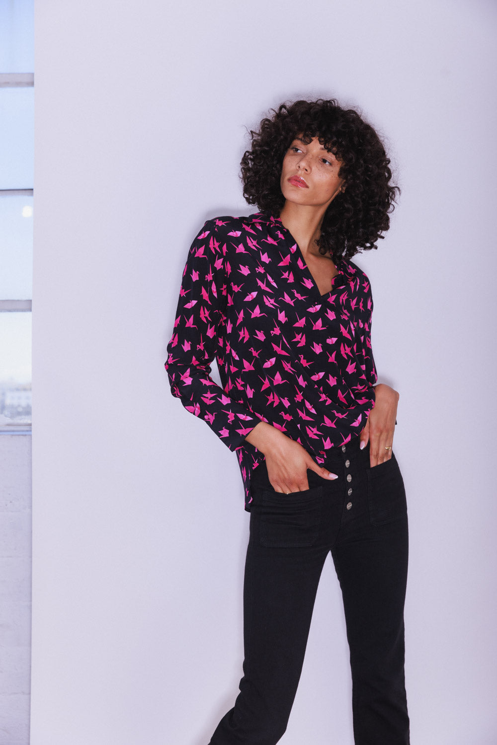 A woman wearing a Catherine Gee Daria French Cuff Silk Pink Crane blouse is black and adorned with a graceful pattern of pink cranes in flight paired with high-waisted black pants that feature buttons on the front, creating a chic and elegant look. The backdrop is a plain white wall with a window on the right side, which subtly highlights the stylish and sophisticated design of the blouse, perfect for adding a touch of elegance to any outfit.