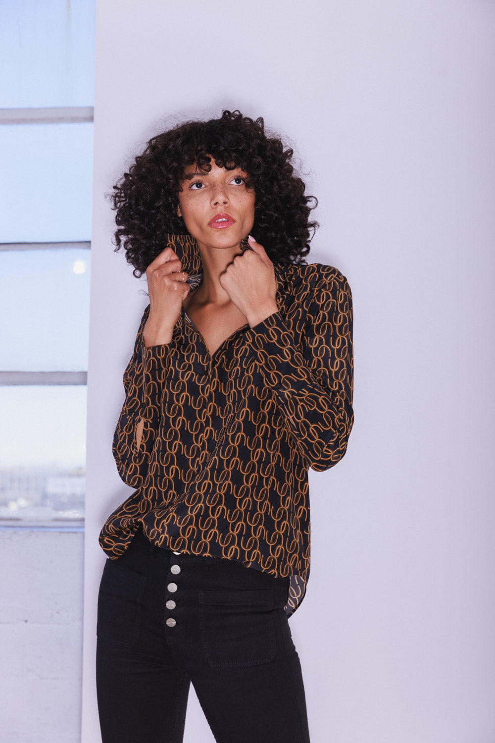 Woman wearing a Catherine Gee Daria French Cuff Silk Blouse CC Print. The blouse is black with a gold chain-link pattern paired with high-waisted black pants with four visible buttons. The background shows a window with a view of a cityscape.