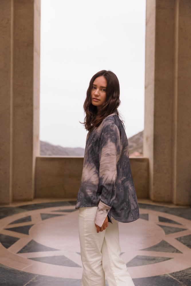 A woman standing in an architectural setting with a circular patterned floor and large stone pillars framing a mountainous background wearing a Catherine Gee Daria French Cuff Silk Smoke blouse features a tie-dye pattern in shades of gray and white. The blouse has a loose fit and long sleeves with French cuffs paired with white pants. The setting and attire create a visually appealing contrast between the structured environment and the fluidity of the blouse's design.