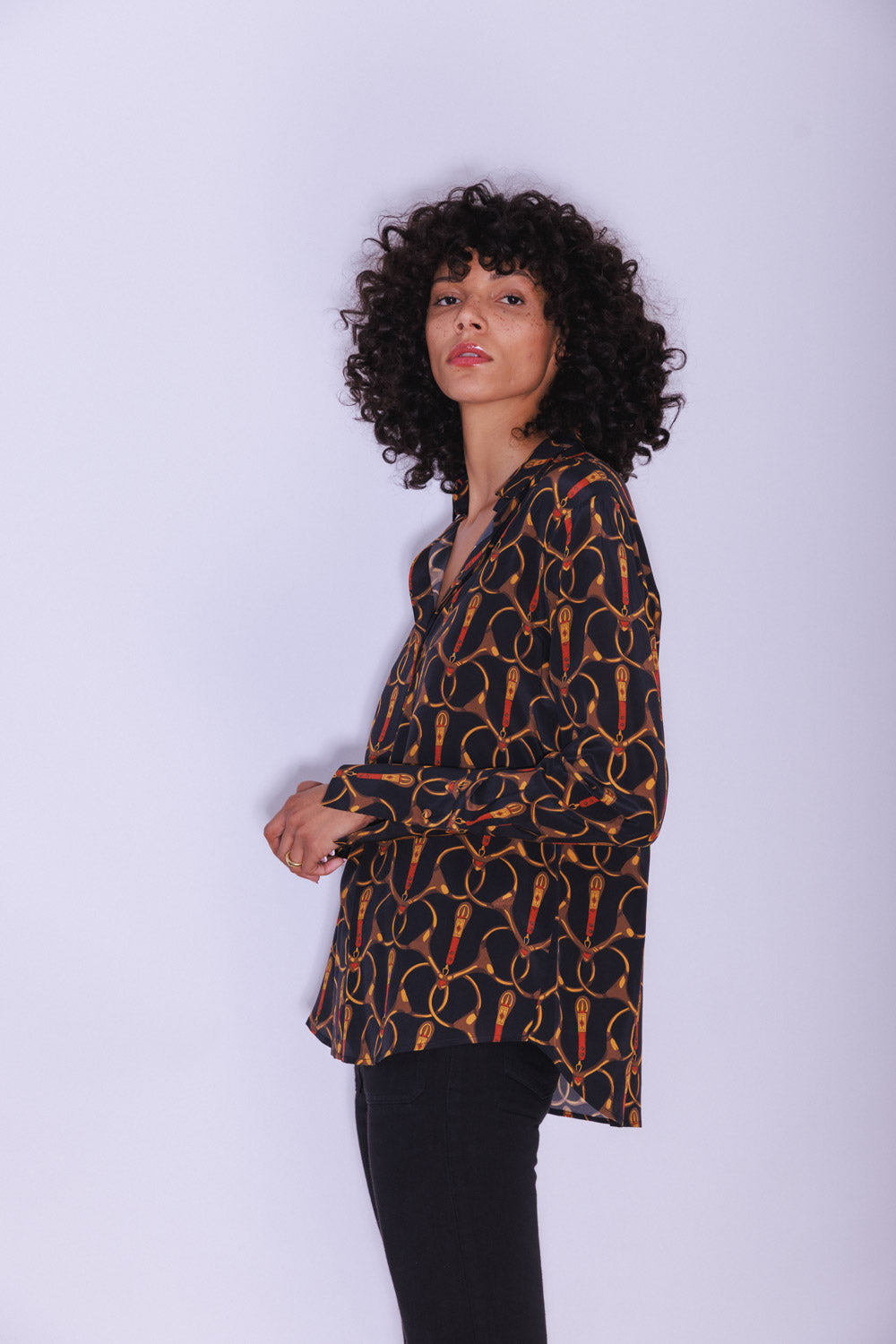 A woman wearing a Catherine Gee Daria French Cuff Silk - Buenos Aires blouse features a dark background with a pattern of intertwined, gold-colored designs resembling ropes or chains. It has a loose fit and long sleeves with French cuffs. She is standing sideways, and they have curly hair on a plain and light-colored background, contrasting with the dark blouse, making the pattern stand out.