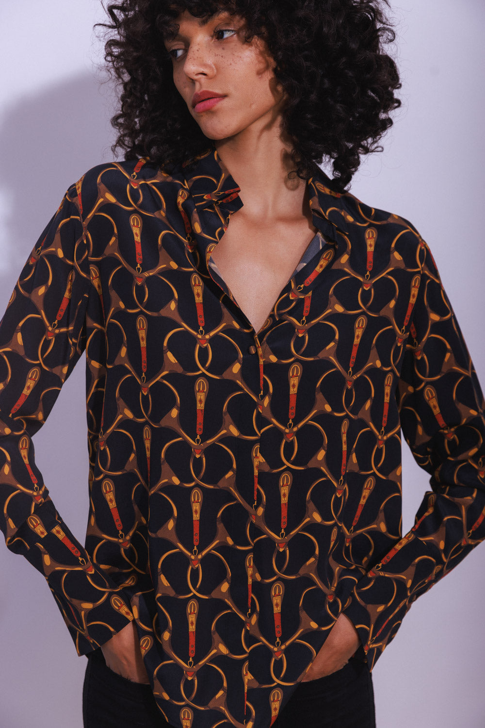 A woman standing against a plain background with her hands in her pockets wearing a Catherine Gee Daria French Cuff Silk - Buenos Aires blouse features a black background with a repeating pattern of gold and red elements resembling stylized horse bits. The blouse has a collar and buttons down the front, with the top button left undone.