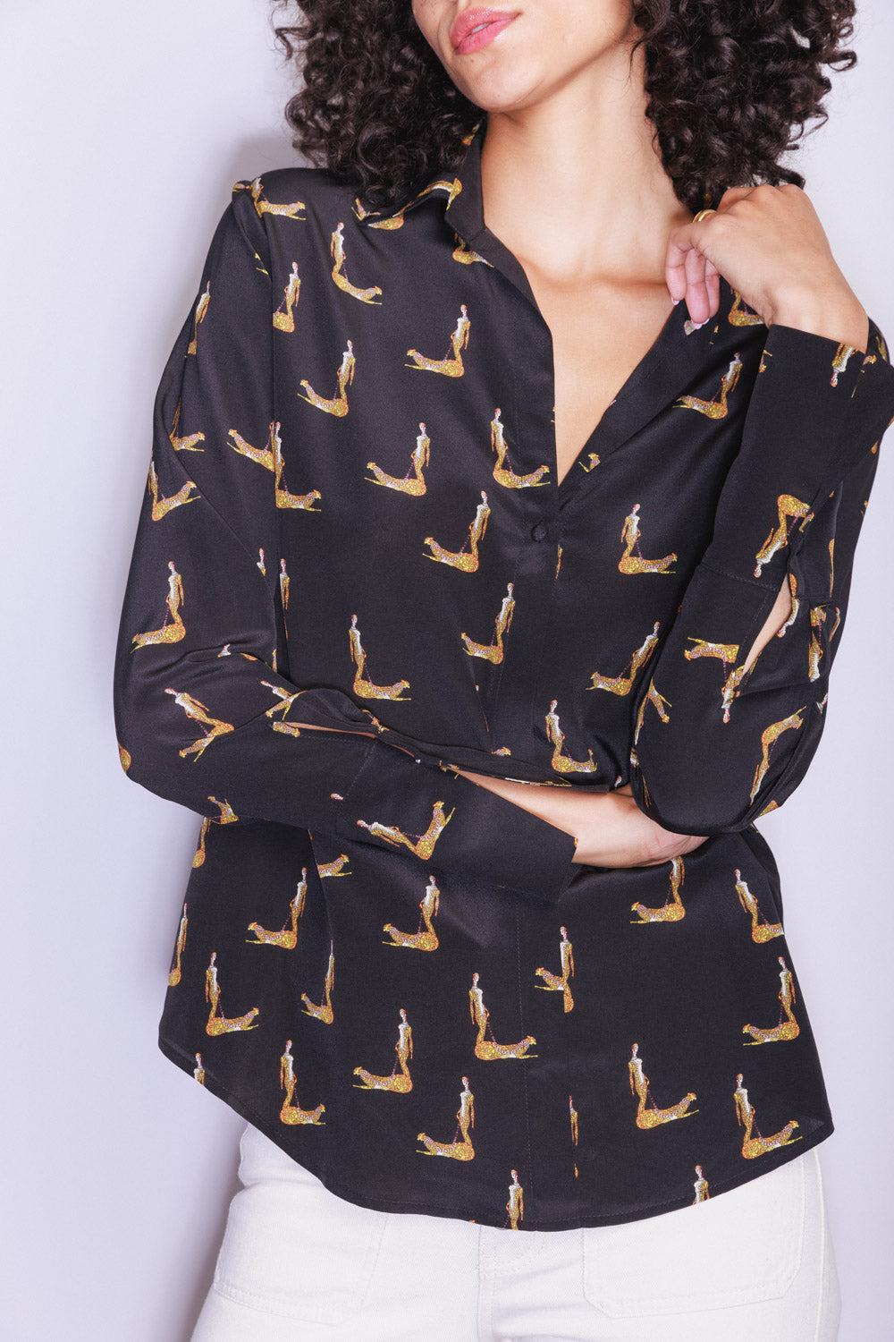 Woman wearing a Catherine Gee Daria French Cuff Silk Blouse Catwoman. The blouse is black with a pattern of a woman in a cat-like pose, repeated throughout the fabric. She has curly hair and poses with one hand resting on the left shoulder and the other arm under the left elbow. The blouse has a collar and buttons down the front, paired with white pants.