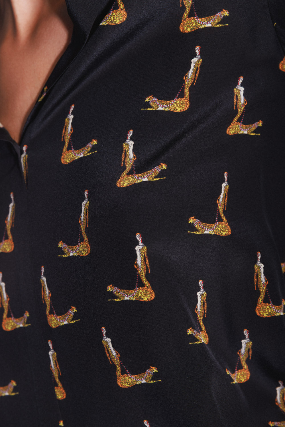A close-up view of a Catherine Gee Daria French Cuff Silk Blouse Catwoman. The blouse is black with repeated images of a stylized Catwoman in a kneeling position, adorned in a glittery gold and red outfit. The fabric appears smooth and shiny, highlighting the intricate and colorful design of the Catwoman figures.