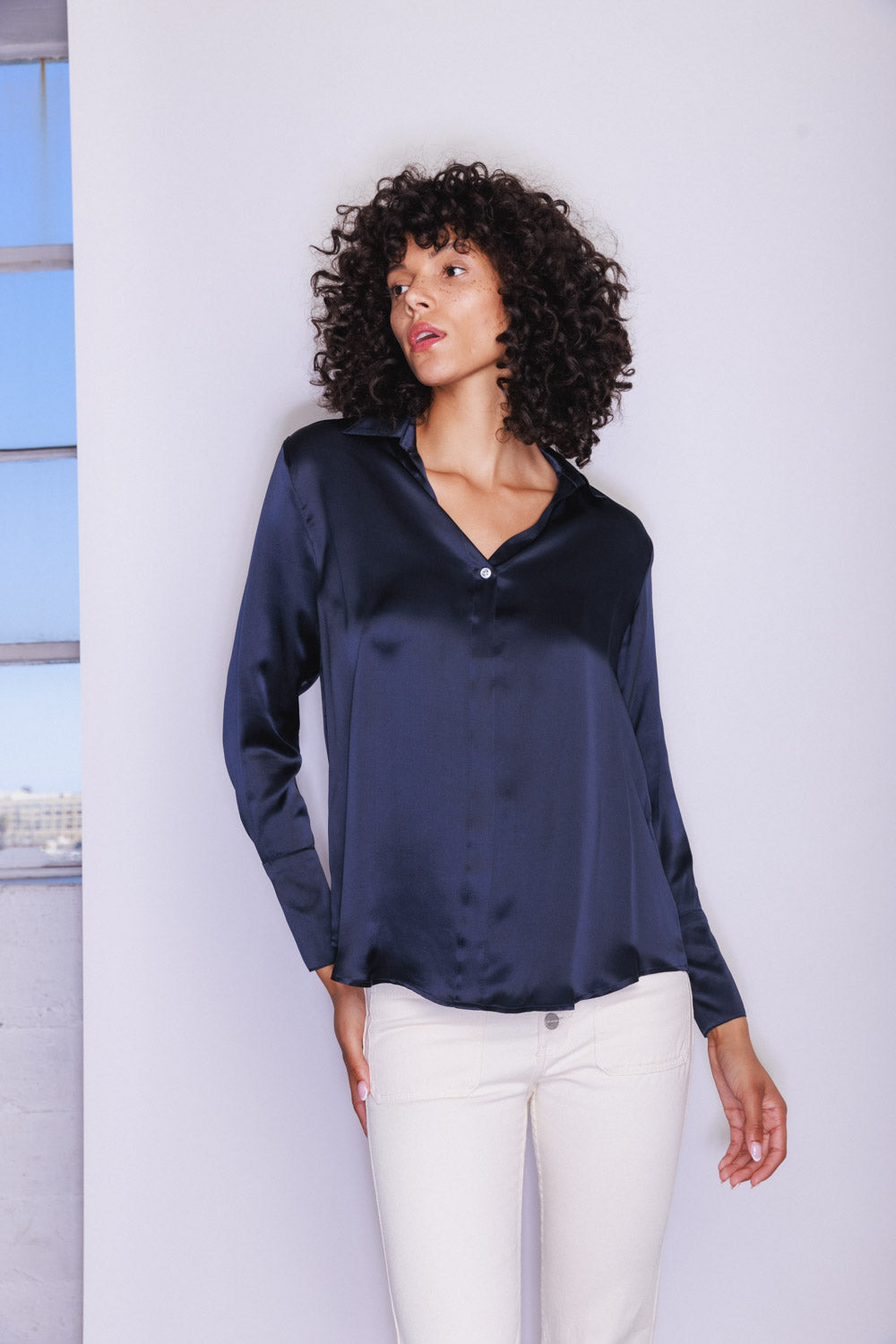 A woman wearing a Catherine Gee Daria French Cuff Silk-Midnight Navy blouse features a smooth, shiny texture and a V-neck with a button closure paired with white colored pants. The background includes a partial view of a window with a blue sky visible outside.