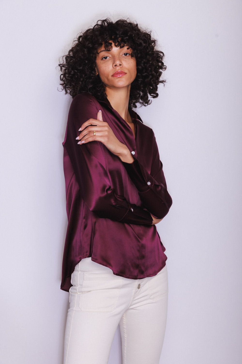 A woman wearing a Catherine Gee Daria French Cuff Silk Wine blouse in a deep wine color with a silky texture, featuring long sleeves and French cuffs paired with white-colored pants, which contrast with the rich hue of the blouse. The blouse has a relaxed fit with a slightly curved hemline, adding to its elegant and sophisticated appearance.