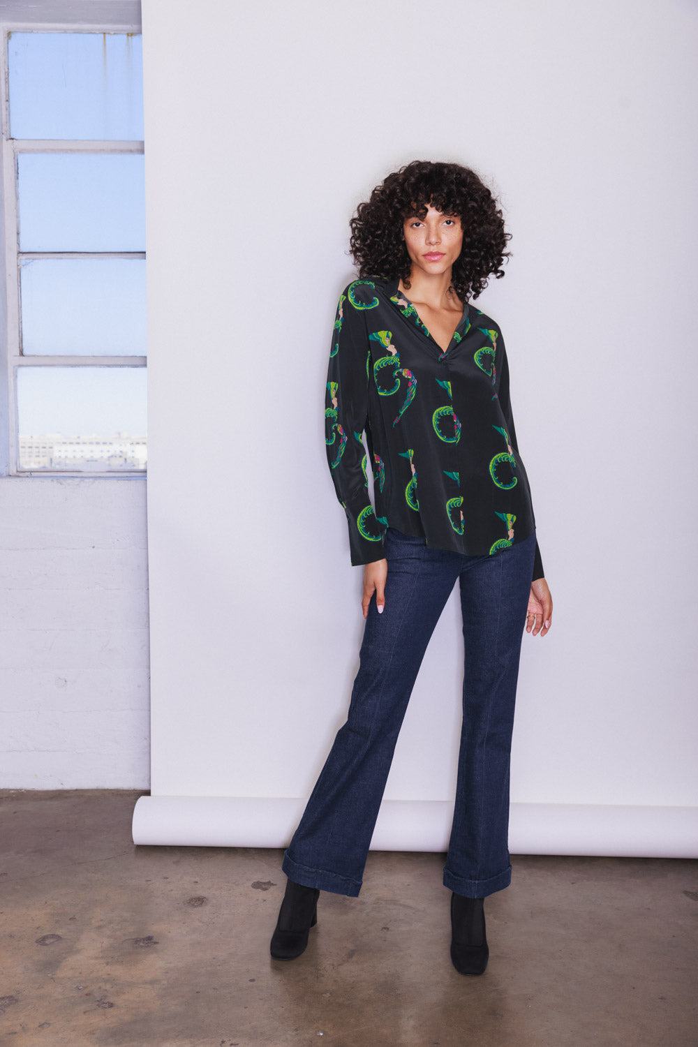 A woman wearing a Catherine Gee Daria French Cuff Silk Blouse Egyptian Goddess. The blouse is color black with a green, blue, red, and pink colors circular pattern, featuring a V-neck and long sleeves with French cuffs paired with dark blue flared jeans and black ankle boots. She is standing against a plain white backdrop with a concrete floor and a window with multiple panes on the left side.