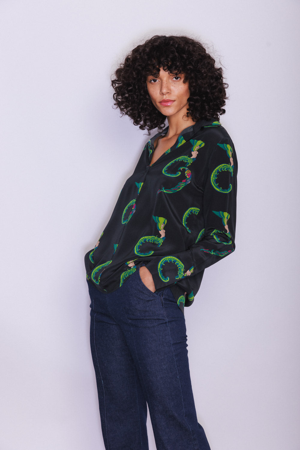 A woman wearing a Catherine Gee Daria French Cuff Silk-Egyptian Goddess blouse features black with green, blue, red, and pink circular pattern designs featuring Egyptian motifs. She is also wearing dark blue jeans and has curly hair on a plain white background.