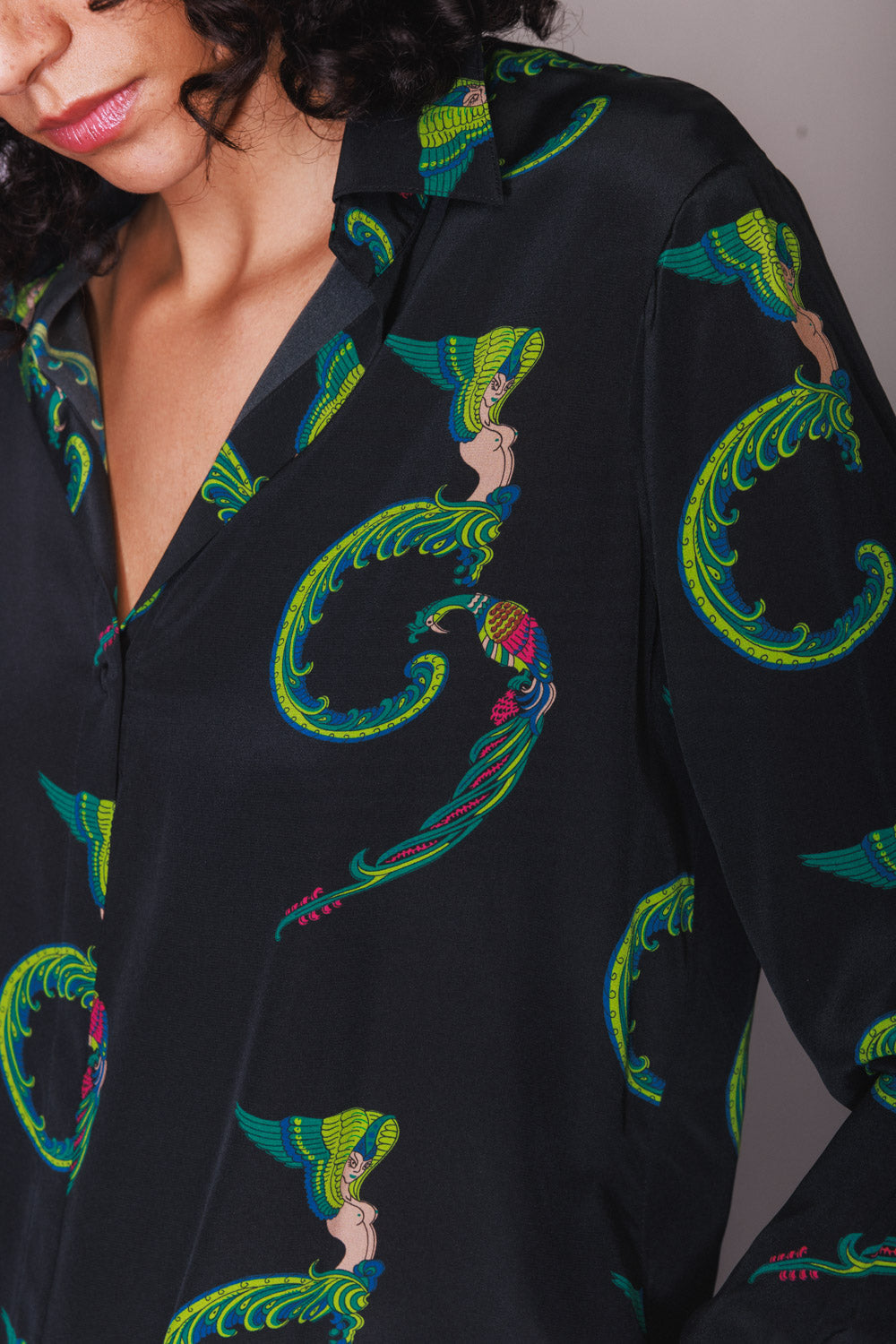 A close-up view of a woman wearing a Catherine Gee Daria French Cuff Silk Blouse Egyptian Goddess. The blouse is black with a vibrant pattern featuring Egyptian goddess motifs in green, blue, red, and pink colors. The design includes stylized figures with elaborate headdresses and flowing garments, creating an elegant and artistic look. The blouse has a collar and a deep V-neckline.