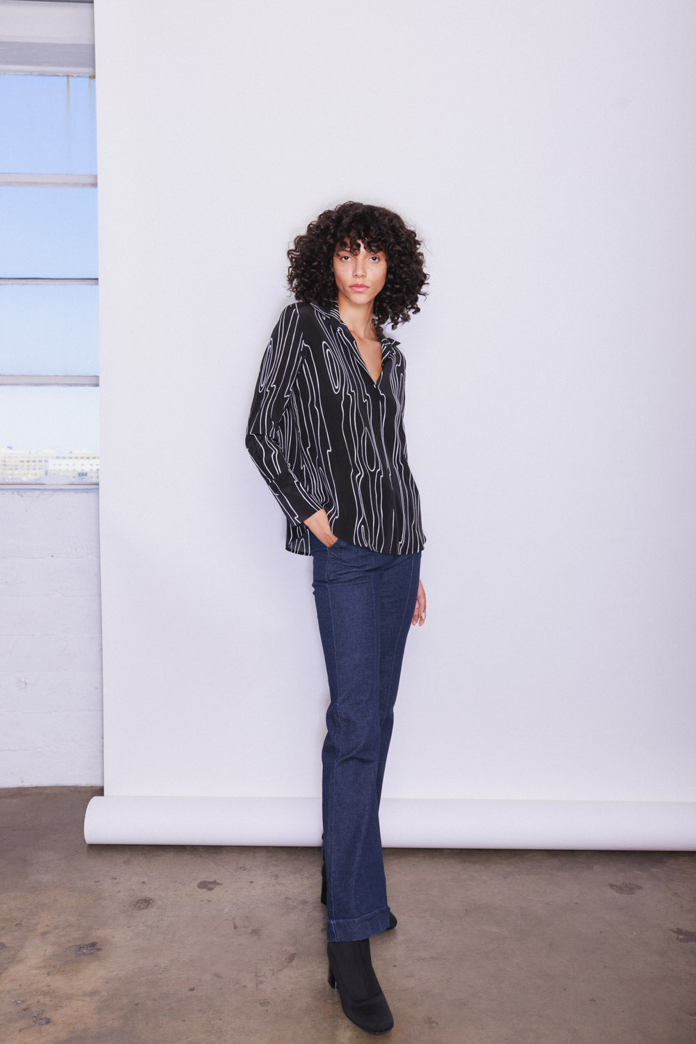 A woman standing against a plain white backdrop wearing a black blouse with white abstract lines, known as the Catherine Gee Daria French Cuff Silk-Byrne paired with blue jeans and black boots showcasing the blouse, poses her hands in the pockets of her jeans, and standing on a concrete floor.