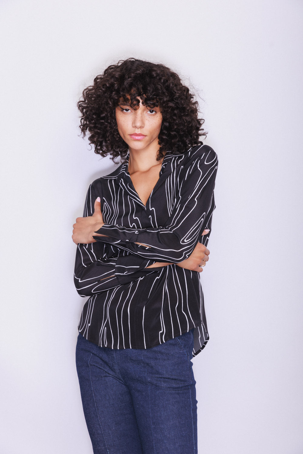 A woman wearing a Catherine Gee Daria French Cuff Silk-Byrne blouse features black with white vertical stripes French cuffs paired with dark blue jeans. The blouse has a relaxed fit and is styled with the sleeves partially covering the hands.