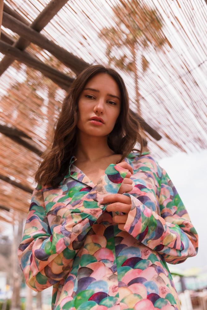 A woman wearing a colorful Catherine Gee Daria French Cuff Silk-Lima blouse features a pattern of overlapping semi-circles in various pastel shades like pink, blue, green, and purple. The blouse has French cuffs and a smooth, shiny texture. She is standing under a wooden pergola with trees and sky visible through the slats.