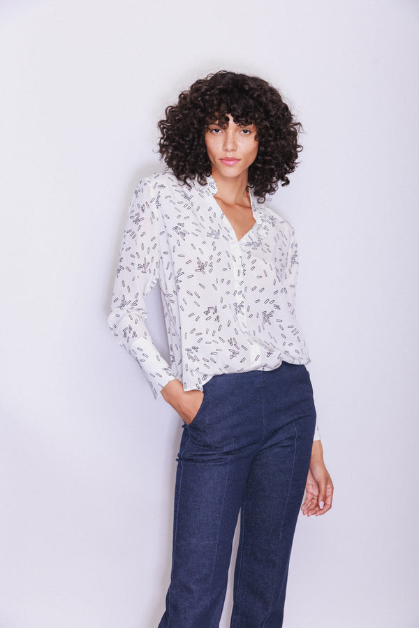 A woman wearing a Catherine Gee Daria French Cuff Silk Pin blouse features white with a delicate pattern of small, scattered prints, a classic collar, and French cuffs, which add a touch of sophistication. The blouse is tucked into a pair of high-waisted, dark blue pants. The overall look is elegant and stylish, suitable for both professional and casual settings.