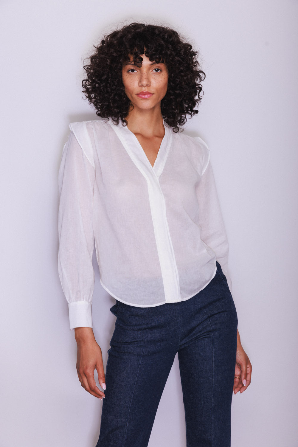 A woman wearing the Catherine Gee Franka White Blouse. This semi-sheer blouse features a high V-neckline with a button-down front, giving it a polished look. The long sleeves end in wide cuffs, adding to its chic design, has a relaxed, loose fit, paired with dark-colored pants, creating a sophisticated and versatile style suitable for both casual and professional settings.