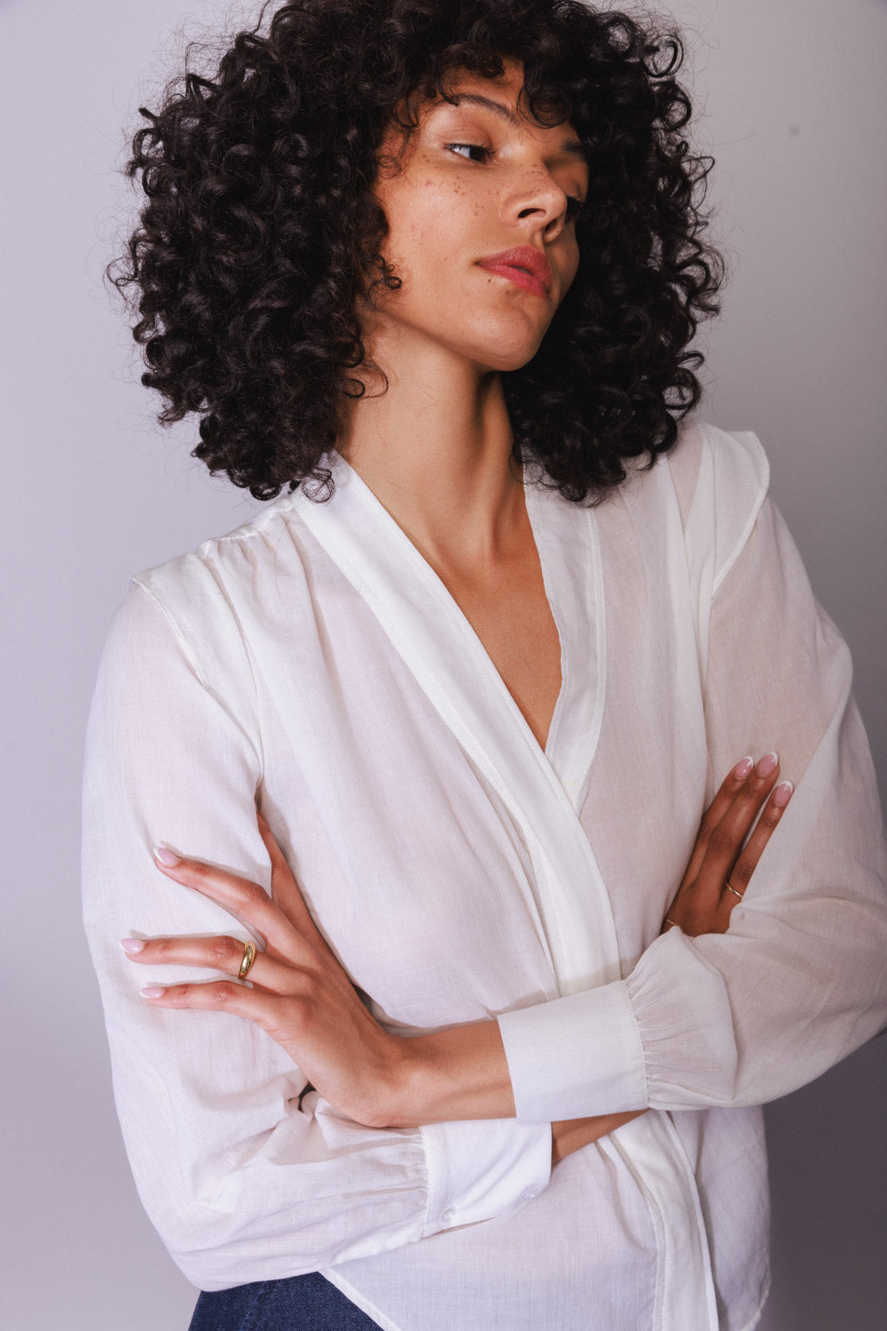 A woman with arms crossed wearing the Catherine Gee Franka White blouse is made of elegant, lightweight fabric with a wrap-style front and a V-neckline. It has long, slightly puffed sleeves that end in buttoned cuffs and appears semi-sheer, revealing a hint of the skin underneath creating a sophisticated and stylish look that's perfect for both professional and casual settings.