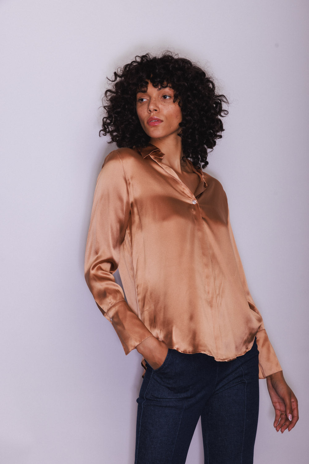 A woman wearing a Catherine Gee Daria French Cuff Silk Bronze blouse features a shiny, smooth texture and a relaxed fit, with French cuffs and long sleeves. She is also wearing dark jeans and standing against a light-colored background.