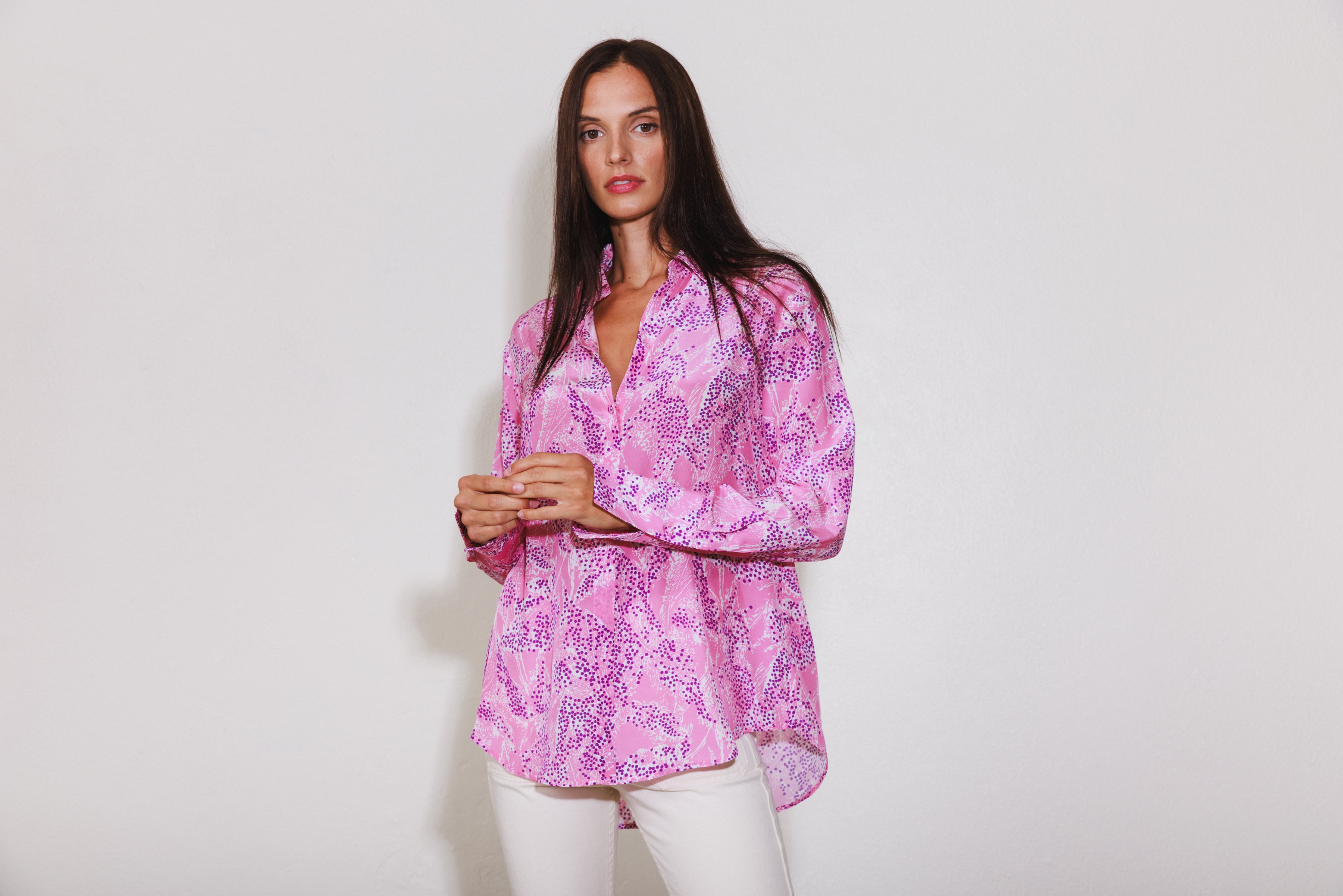 A woman wearing a Catherine Gee Daria French Cuff Silk Pink Sea blouse is predominantly pink with a pattern resembling abstract sea elements in darker shades of purple and white. It has a classic collar, long sleeves with French cuffs, and a high-low hemline paired with white pants, creating a chic and sophisticated ensemble. The fabric appears to be lightweight and silky, giving it a luxurious and elegant look. 