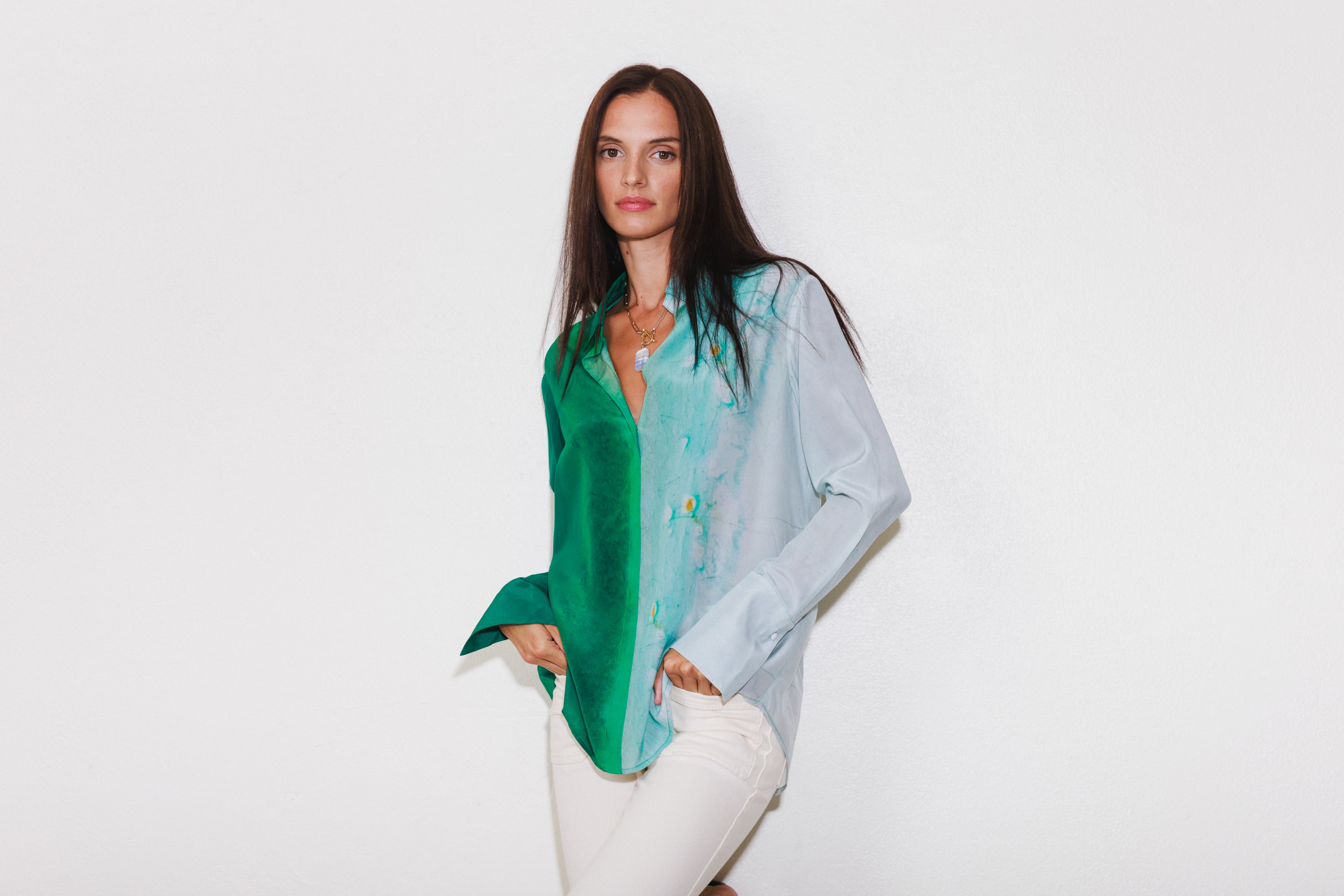 A woman wearing a Catherine Gee Daria French Cuff Silk-King Wash blouse, featuring a blend of green and white colors. The blouse has a classic collar and long sleeves with French cuffs paired with white pants and a necklace. The background is plain white, highlighting the colors and design of the blouse.