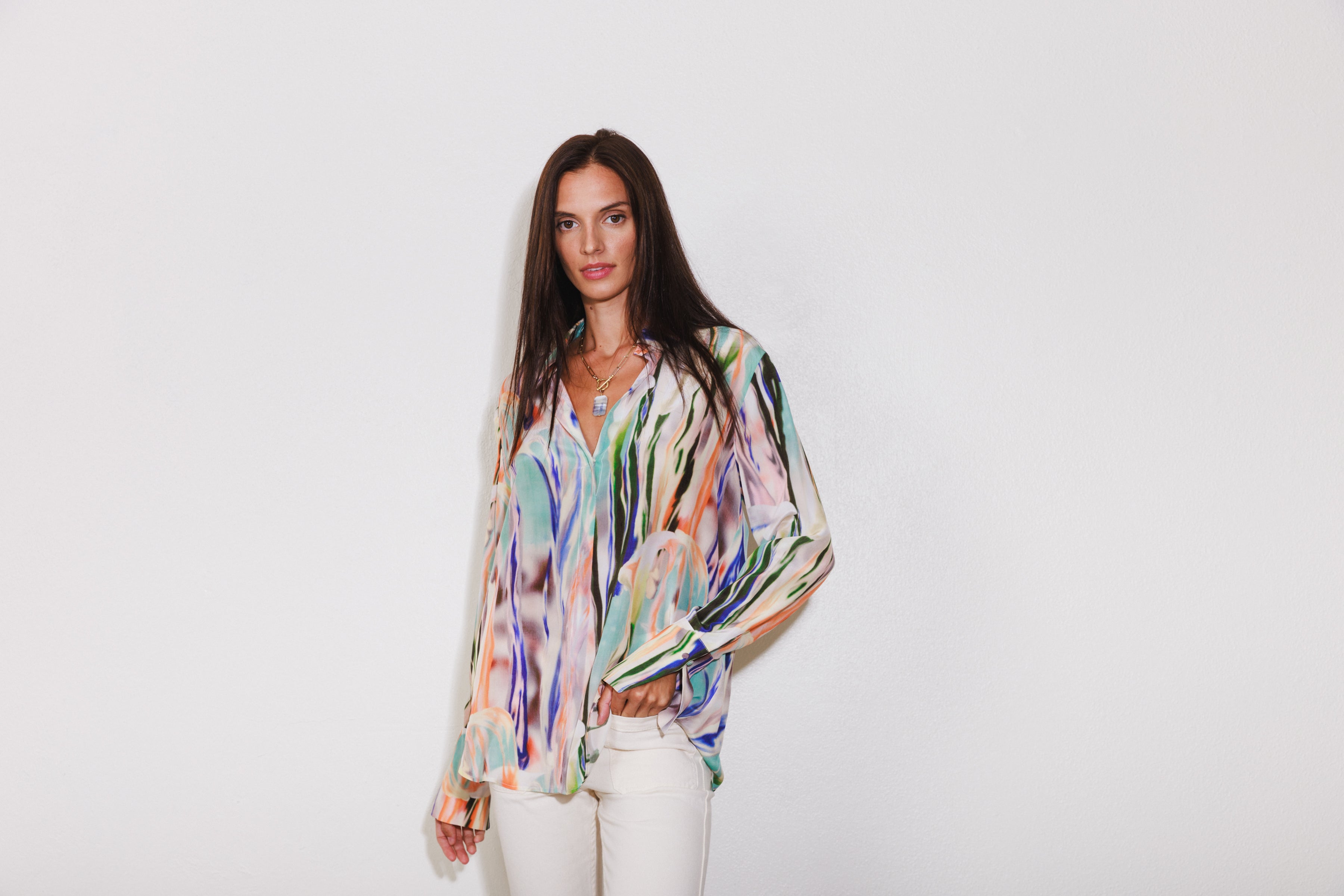 A woman wearing a Catherine Gee Daria French Cuff Silk Abstract Orchid blouse features a vibrant, multicolored abstract design with shades of green, blue, orange, pink, and black paired with white pants and has long, straight hair. The blouse has a loose fit with long sleeves and French cuffs. 
