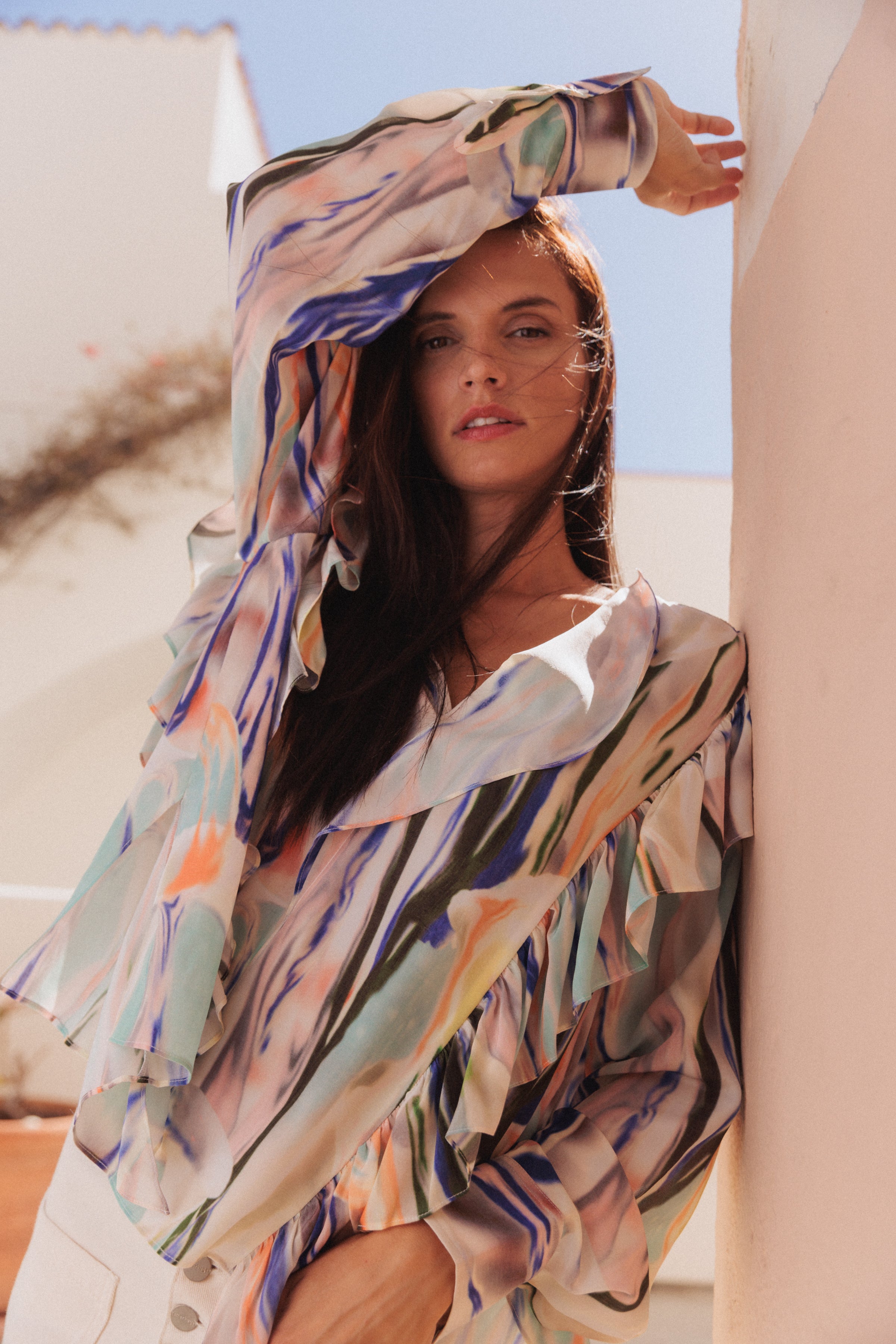 A woman is wearing a Catherine Gee Bella Blouse - Abstract Orchid posing outdoors leaning against a wall with one arm raised above their head. The blouse features a colorful, abstract floral design with shades of blue, green, orange, and white. The blouse has ruffled details on the sleeves and a V-neckline. The background shows a clear sky and part of a building.