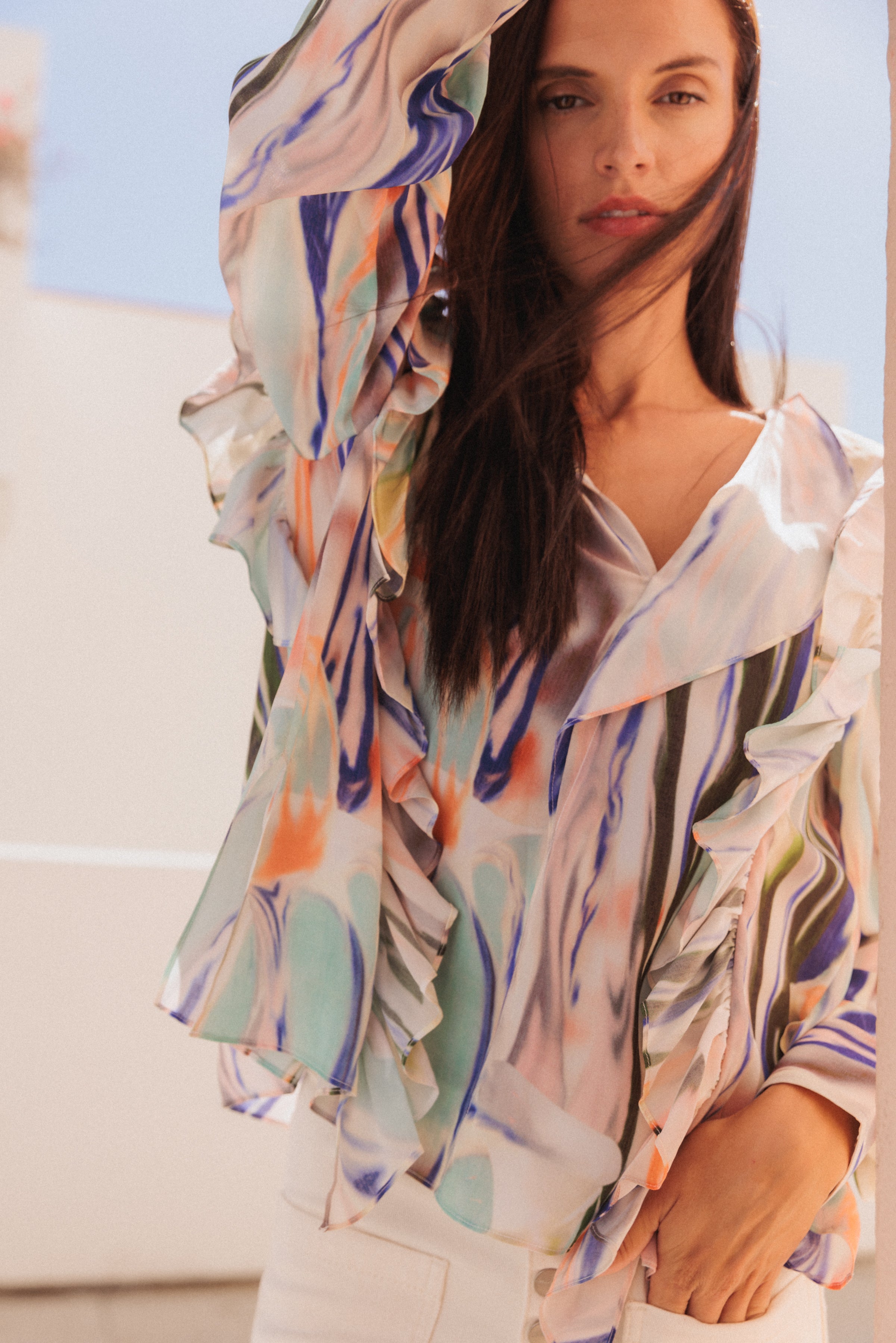 A woman wearing a Catherine Gee Bella - Abstract Orchid blouse features a colorful, abstract design with shades of blue, green, orange, and white, and has ruffled details along the sleeves and front paired with white pants. She is posing with one arm raised in a light, outdoor background setting with a clear sky.