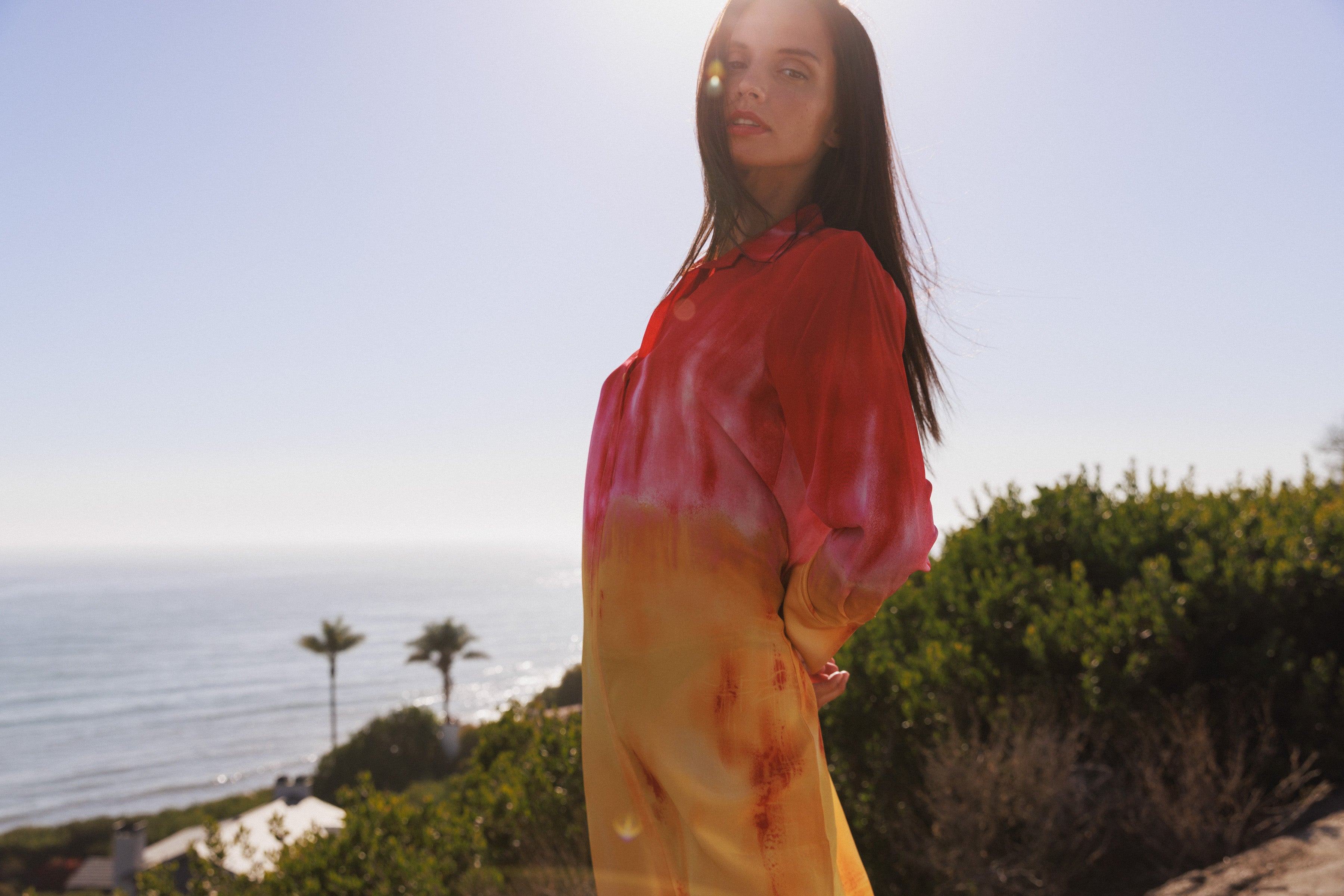 A woman wearing a Catherine Gee Daria Silk Shirt Santa Barbara Sunset dress features a stunning gradient design, transitioning from red and pink hues on the top to yellow at the bottom, capturing the essence of a sunset. It has long sleeves, a loose, flowing fit, and a collared neckline. The luxurious silk material adds to its elegant appearance. She is standing outdoors with a scenic ocean view, palm trees, and greenery, enhancing the summery appeal of the dress.
