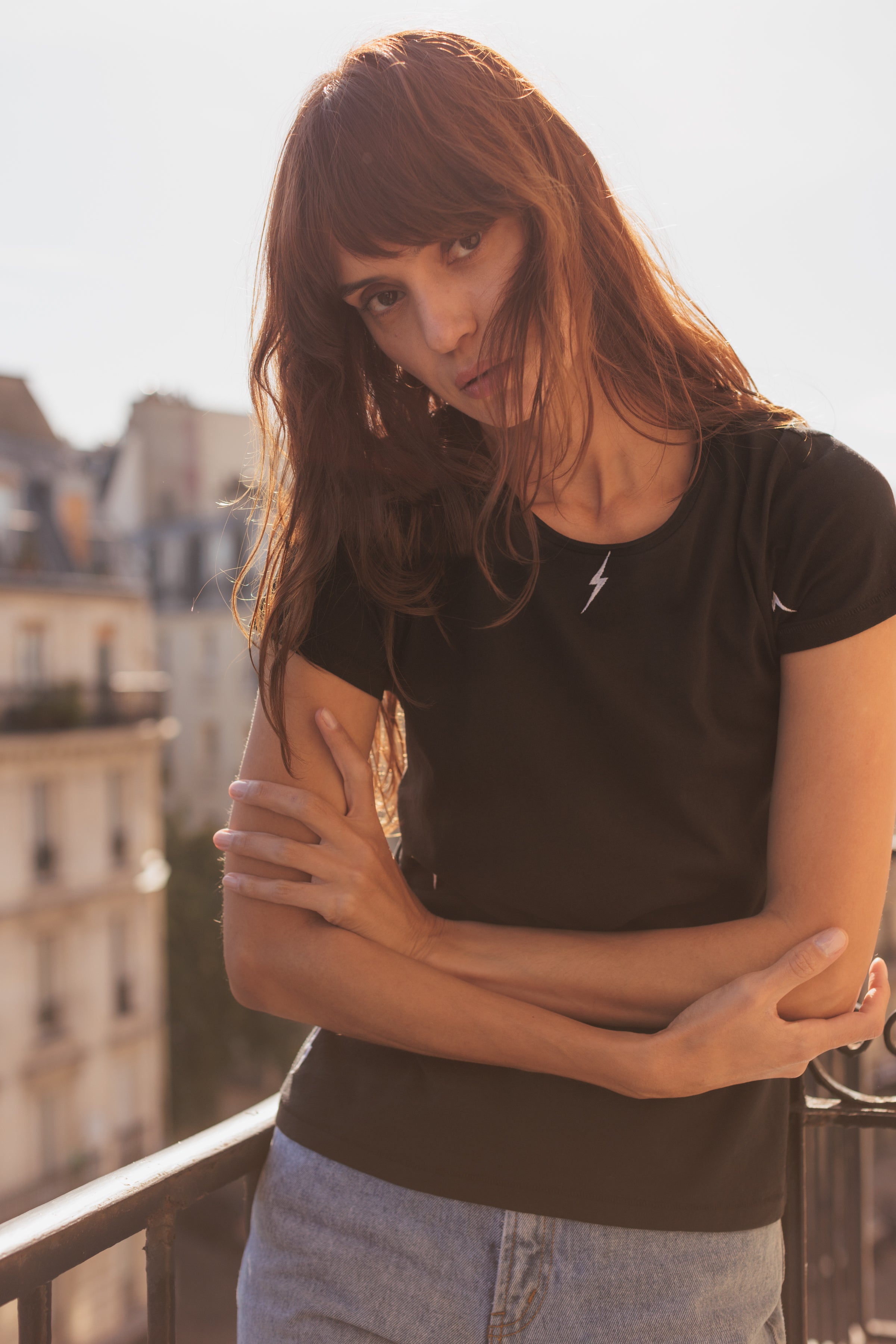 A woman standing on a balcony, wearing a black Catherine Gee Embroidered Cotton Bolt T-Shirt features a small, white lightning bolt embroidered on the chest paired with light blue jeans and has long hair. The background includes a blurred view of buildings, suggesting an urban setting that highlights the casual yet stylish design of the t-shirt, making it interesting for fashion enthusiasts.
